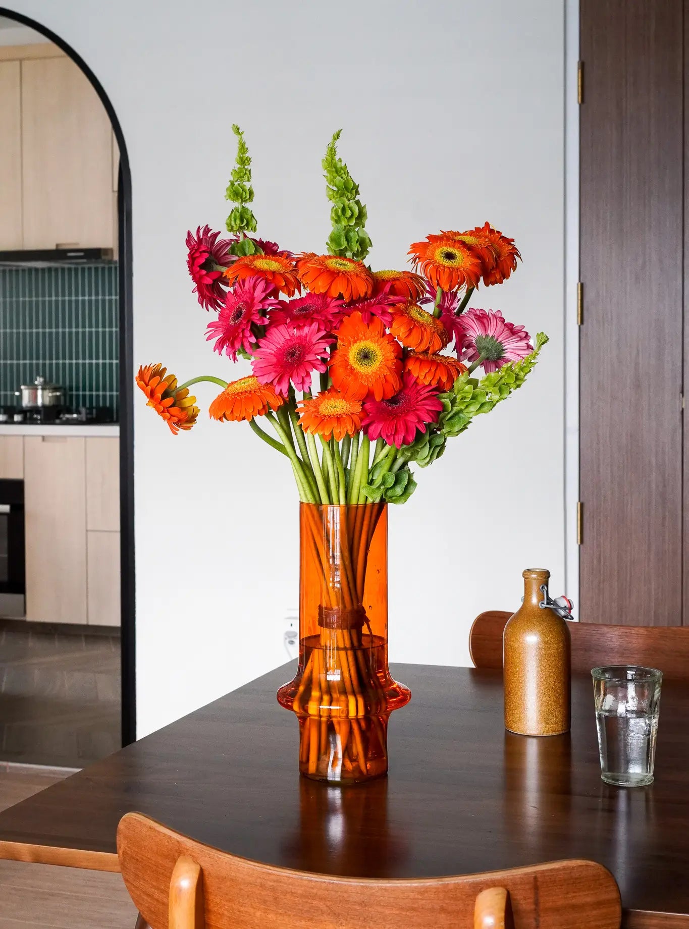 A tall amber glass vase from Wonderland Botanicals Paprika - Wonderland Botanicals Collaboration, filled with a vibrant floral arrangement, stands as a stunning dining table centerpiece amidst a modern kitchen setting.