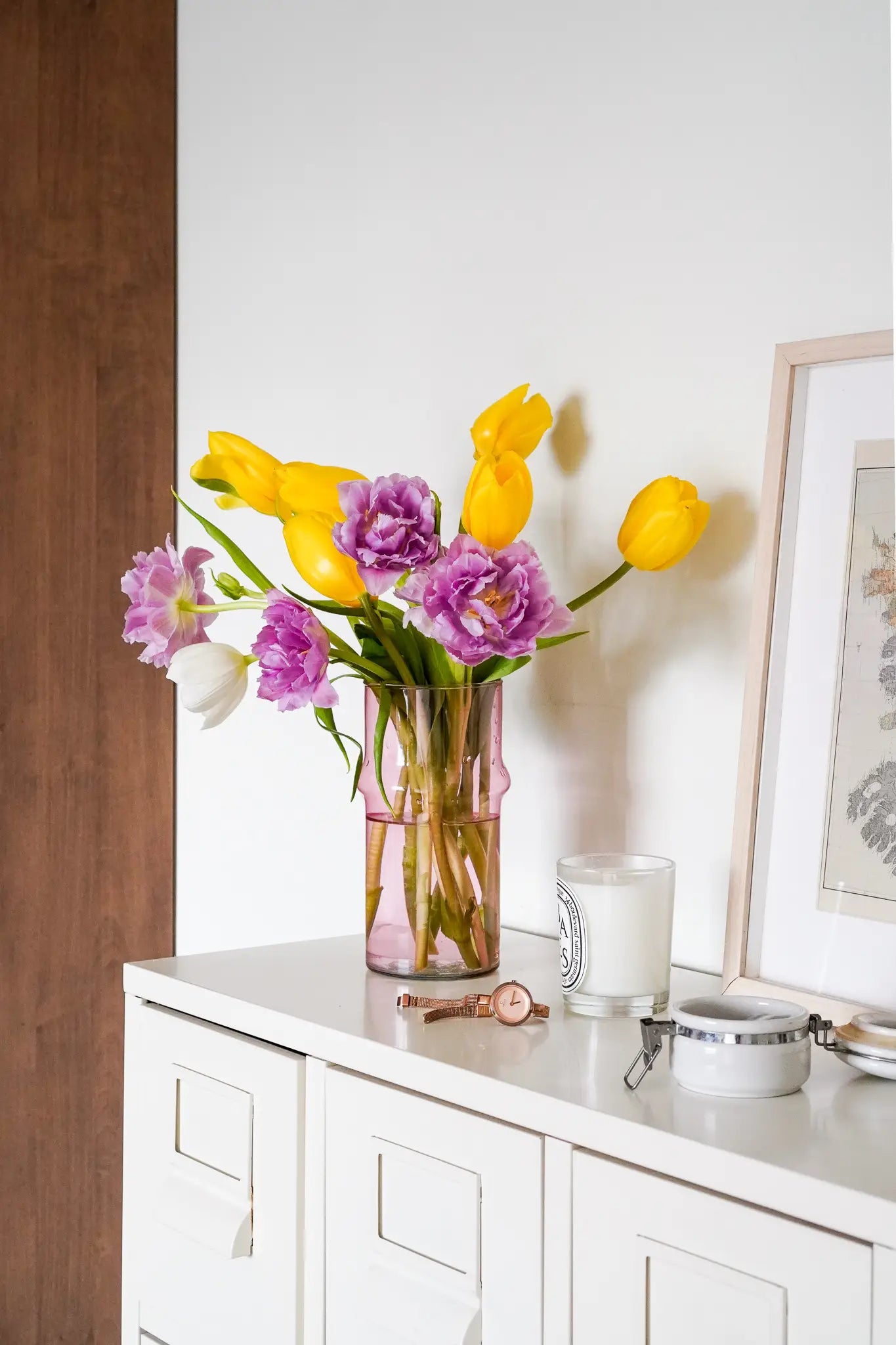 The Rosé vase by Wonderland Botanicals displays yellow tulips, purple flowers, and a white bloom on a white cabinet, ideal for home decor. Nearby: candle, watch, eyeglasses, and framed artwork leaning against the wall.