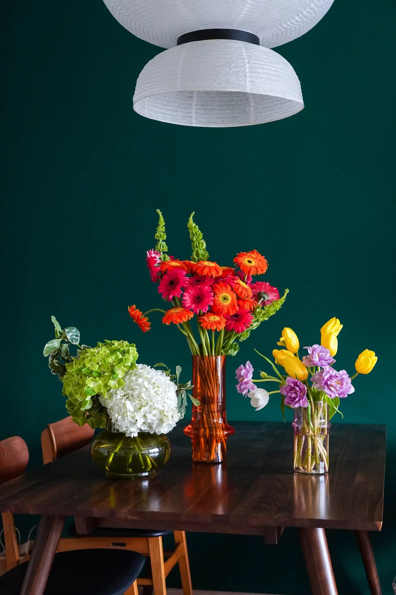 A dark wooden table displays Wonderland Botanicals’ Rosé vase, filled with hydrangeas, gerbera daisies, tulips, and lisianthus. Its rose pink hue adds softness against a deep green wall beneath a white pendant lamp.