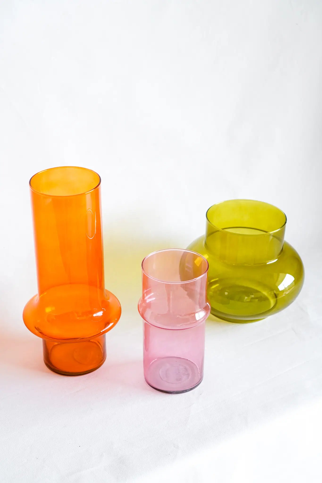 Three modern glass vases from Wonderland Botanicals’ “Rosé - Wonderland Botanicals Collaboration” in orange, rose pink, and green sit on a white surface. The orange and pink are cylindrical; the green is round—perfect for tulips.