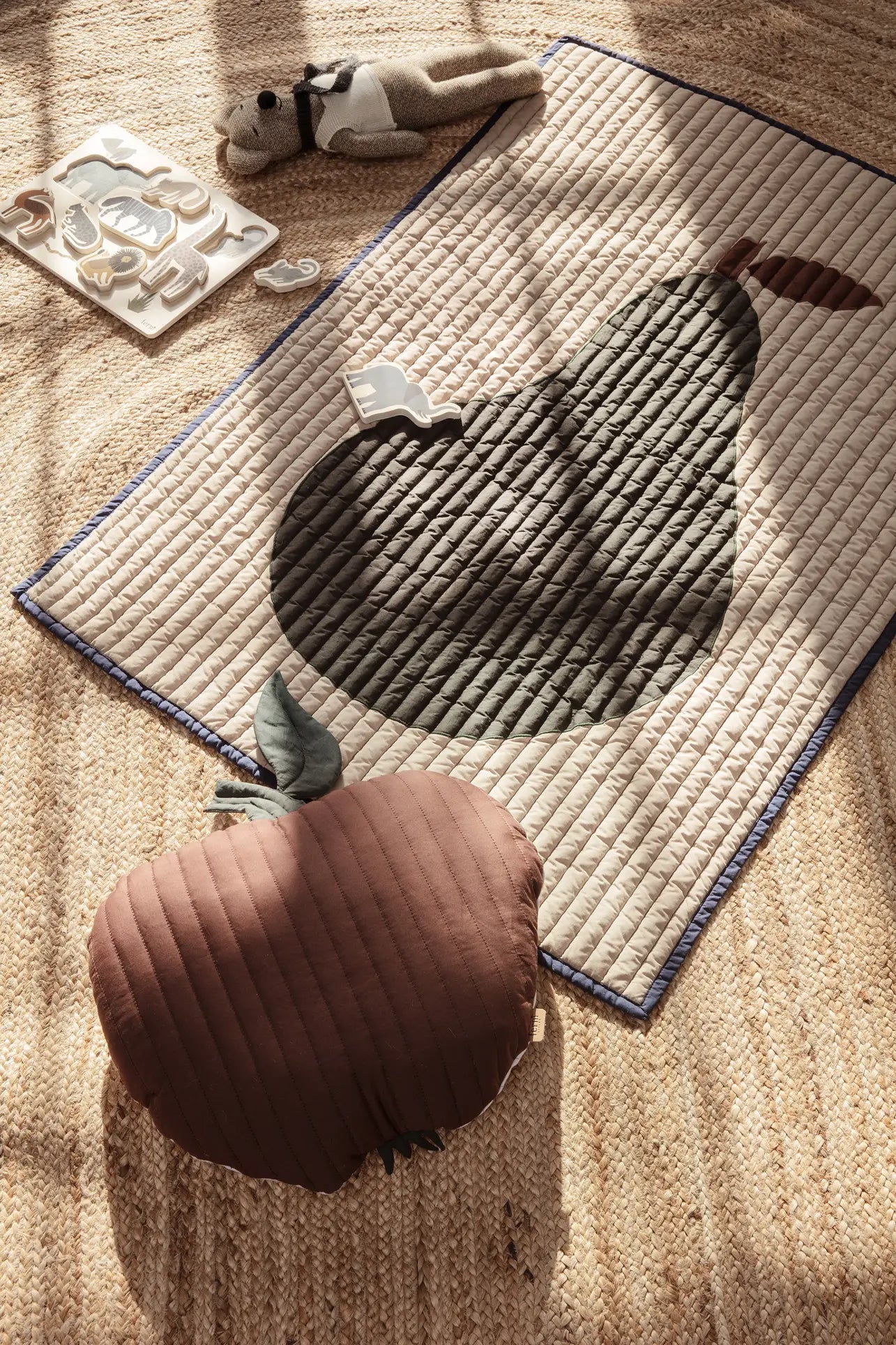 A ferm LIVING Apple Quilted Cushion rests on a woven rug beside a quilted cotton play mat, plush toy, and wooden animal tray in a childs room, with sunlight casting gentle shadows across the scene.