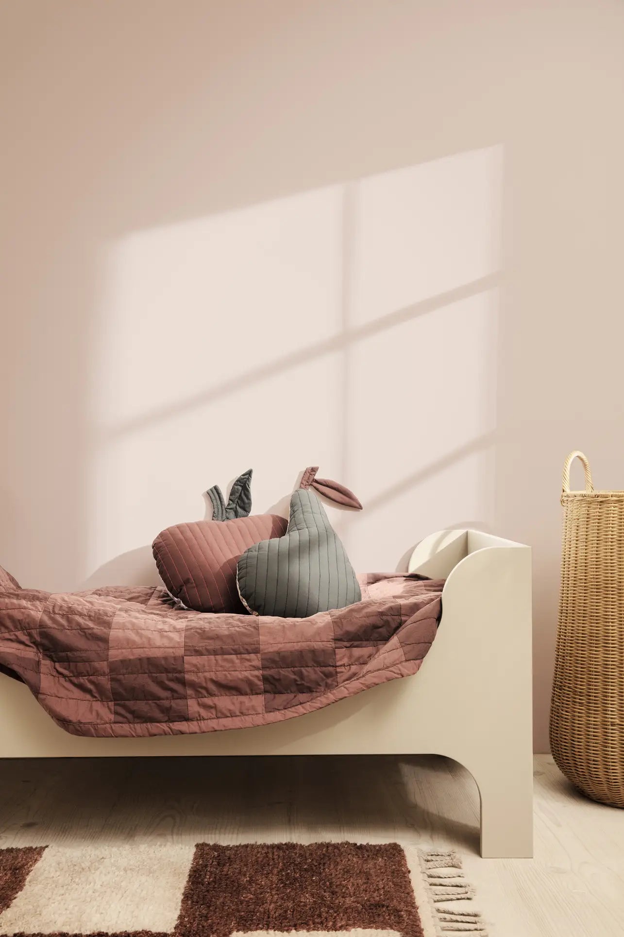 A minimal beige bed with a mauve and brown quilt is in a softly lit child’s room. Two ferm LIVING Apple Quilted Cushions rest on the bed, while a tall woven basket stands nearby. Shadows play across the pale pink wall.