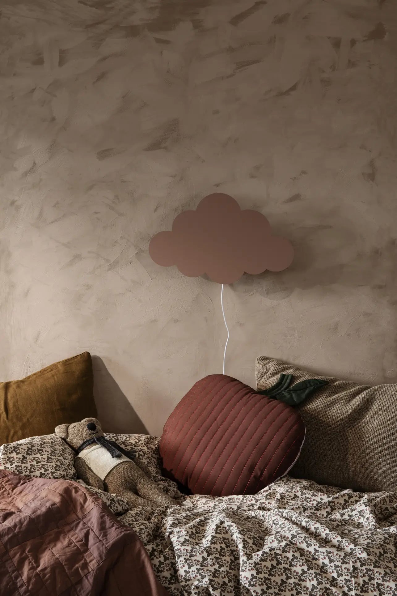 A cozy child’s room with organic quilted cotton bedding, the ferm LIVING Apple Quilted Cushion, brown and red soft cushions, a stuffed bear, and a cloud-shaped wall lamp above the bed against a textured beige wall.