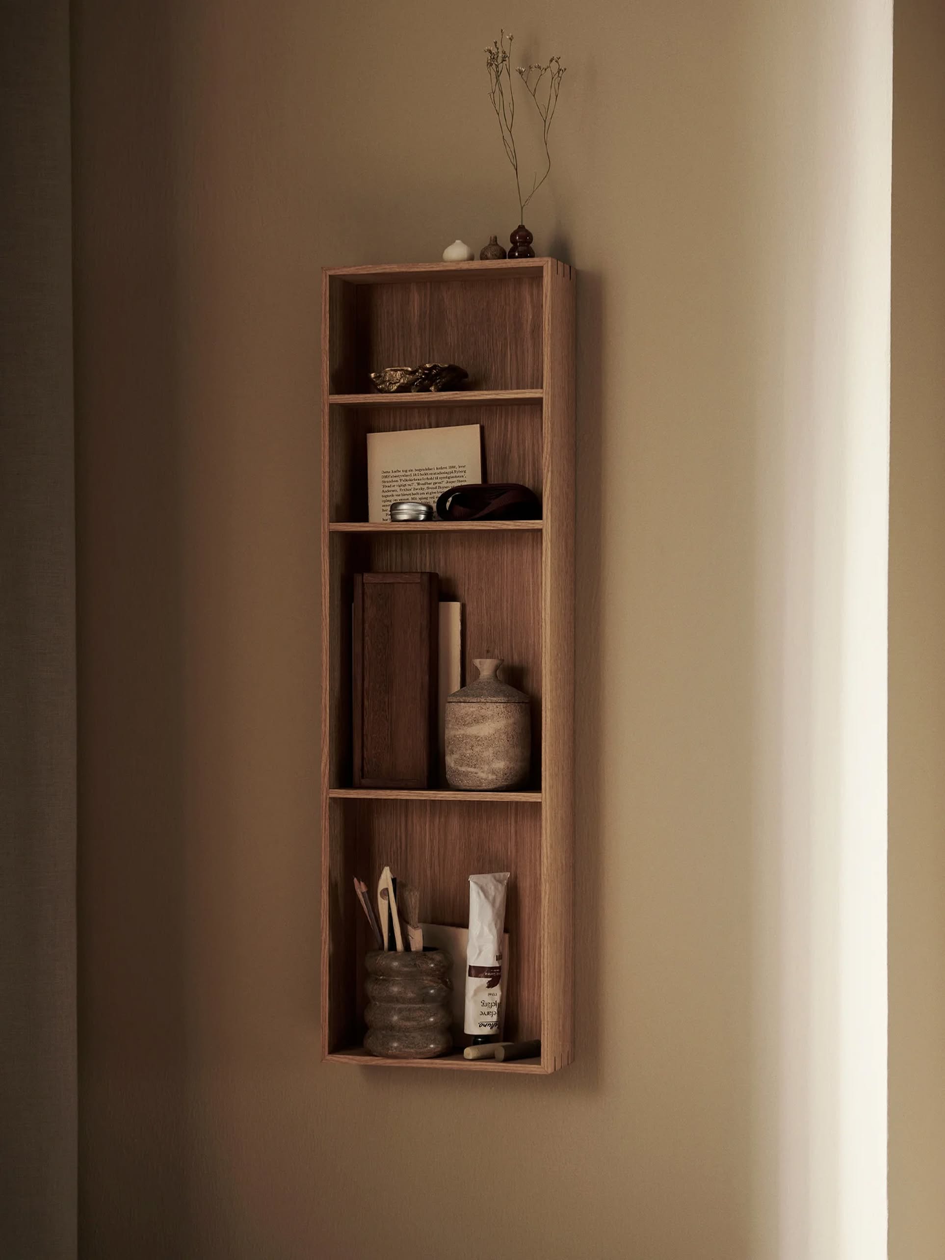 A wooden wall-mounted shelf displays decorative objects, including a ferm LIVING Bendum Cup, a framed photo, books, dried flowers, and a bowl—all elegantly arranged against a beige wall with soft lighting.