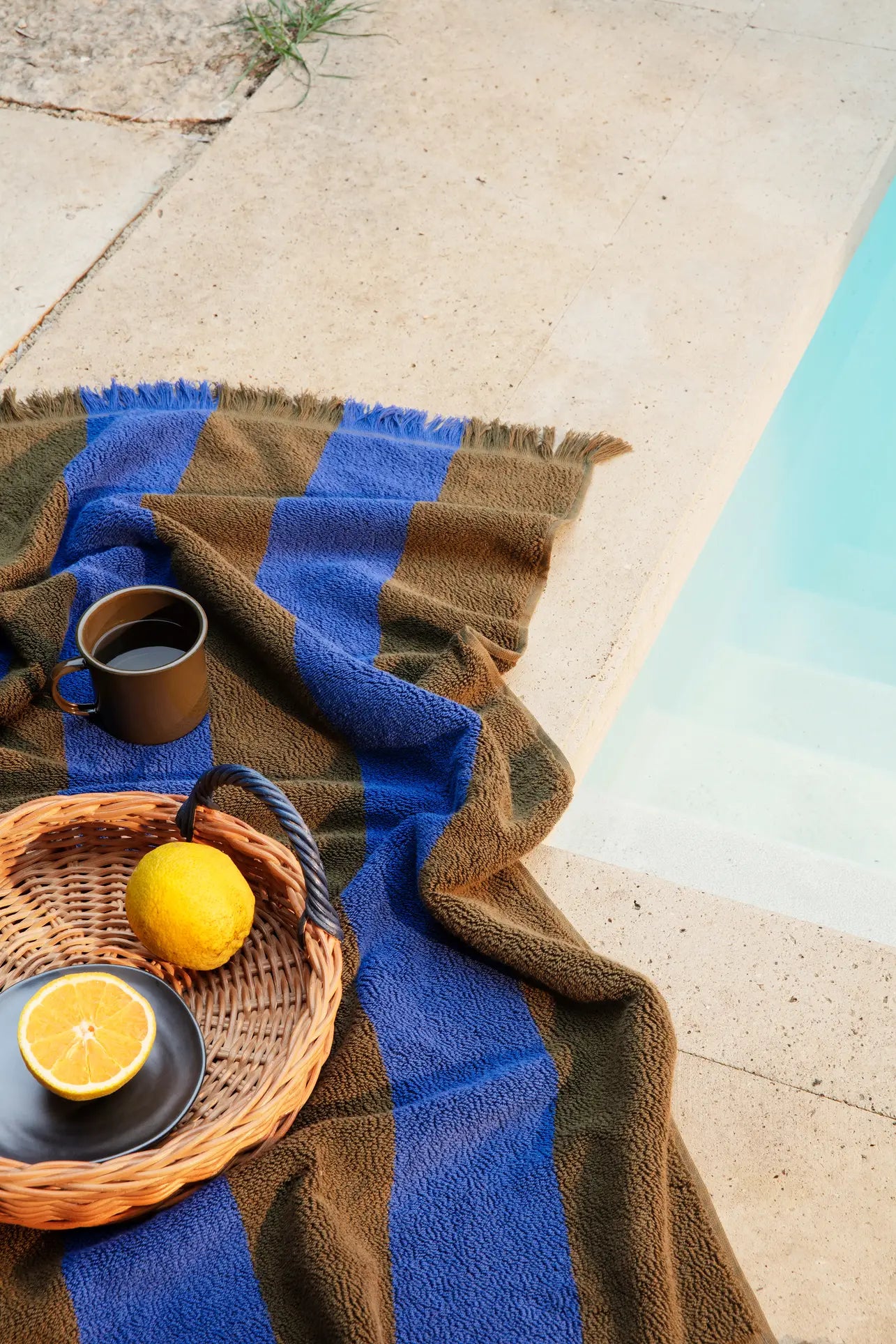 A striped blue and brown towel lies by the pool, with the ferm LIVING Blue Handle Tray from ferm LIVING holding a cup of coffee, a lemon, and a halved orange on a black plate, creating an inviting poolside scene.