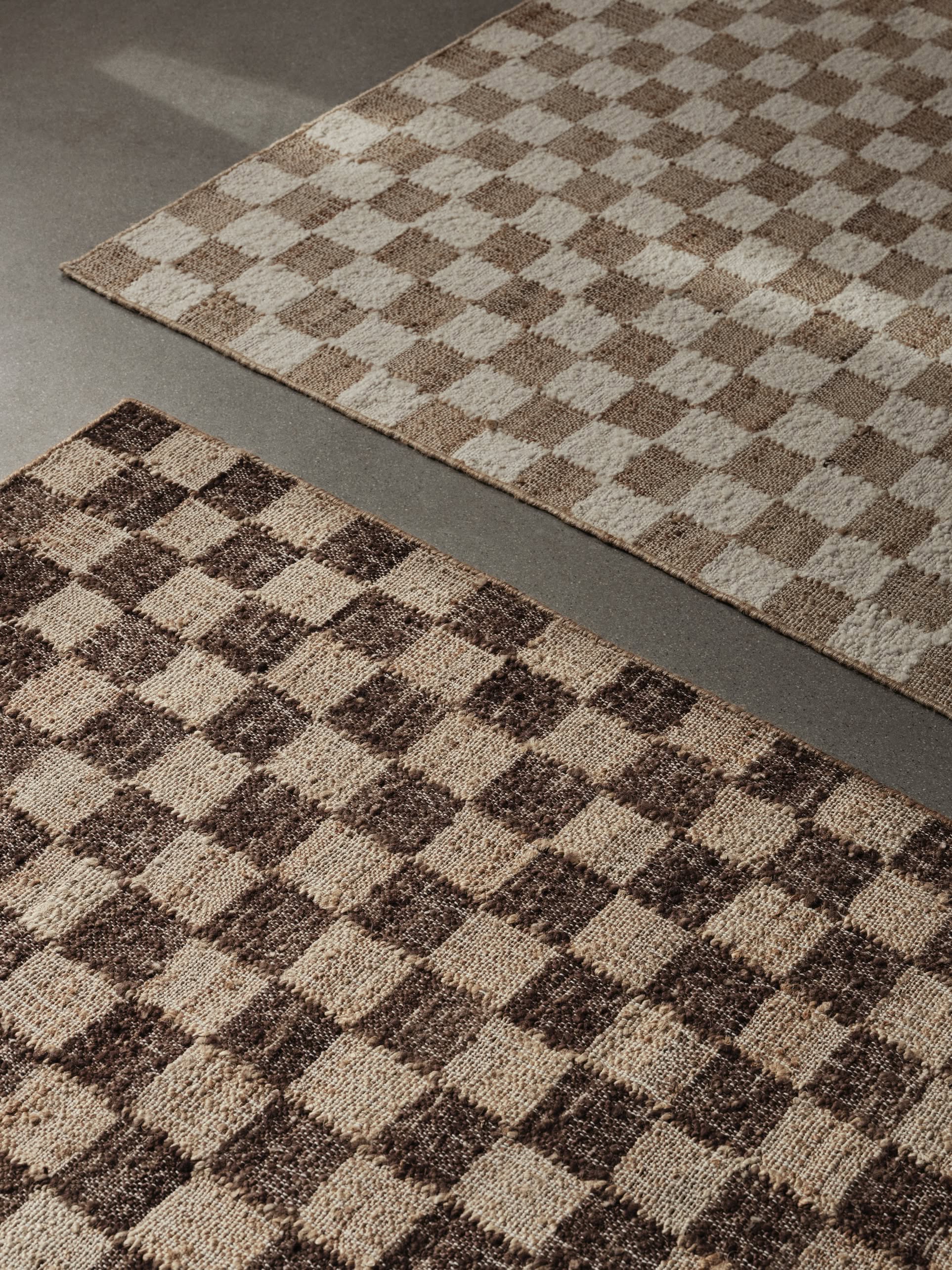 Two ferm LIVING Check Wool Jute Rugs (200x300) with a classic check pattern in neutral tones are on a smooth gray floor; the nearer rug has darker brown squares, while the one farther away displays lighter beige hues.