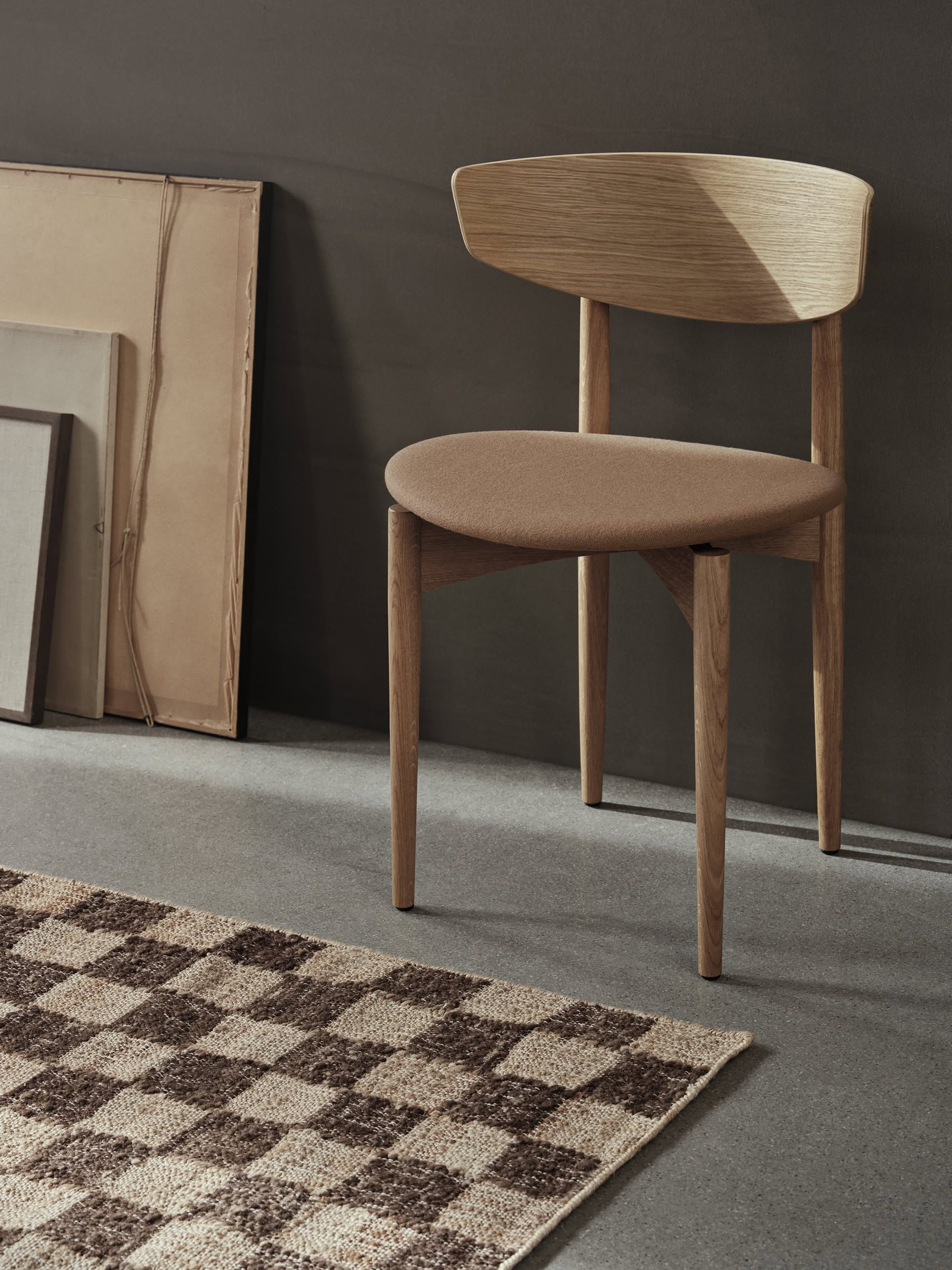 A modern wooden chair with a curved backrest and upholstered seat sits next to framed artwork on a grey floor beside the ferm LIVING Check Wool Jute Rug (200x300), featuring a timeless beige and brown check pattern.