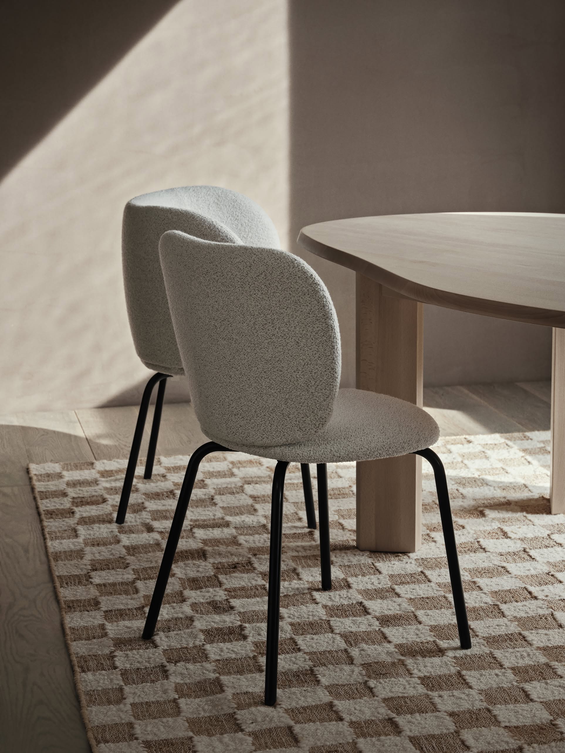 Two modern, light gray upholstered chairs with black legs sit beside a light wood table on a ferm LIVING Check Wool Jute Rug (200x300) by ferm LIVING in a softly lit, minimalist room.