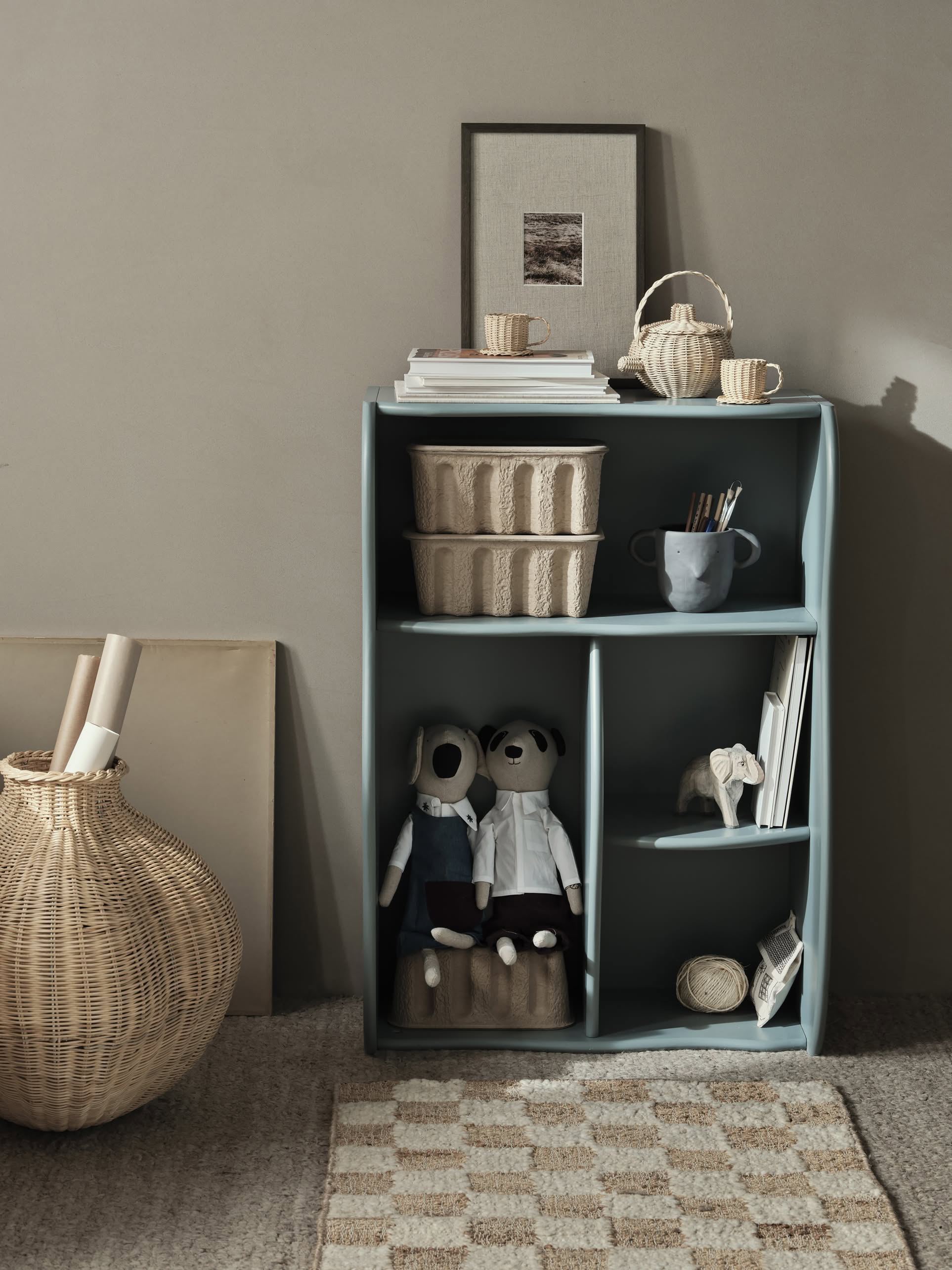 A blue shelf displays baskets, books, a framed picture, mugs, art supplies, plush dolls, and an elephant figurine. Next to it sits a wicker basket with papers on the ferm LIVING Check Wool Jute Rug (200x300) by ferm LIVING.