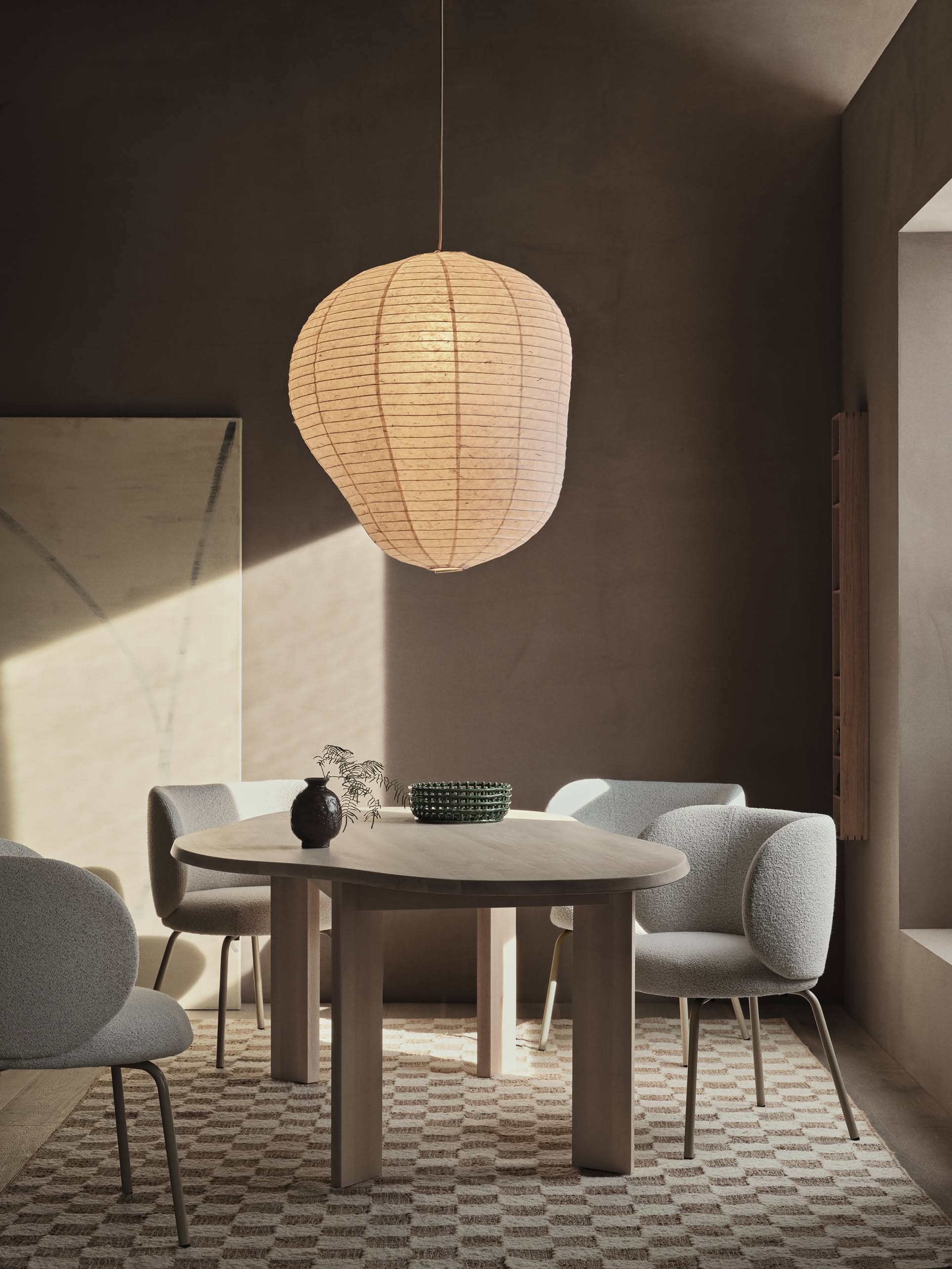 A modern dining area features a round wooden table, four light gray upholstered chairs, a large paper lantern pendant, minimalist decor, and soft natural light streaming over the ferm LIVING Check Wool Jute Rug (200x300).