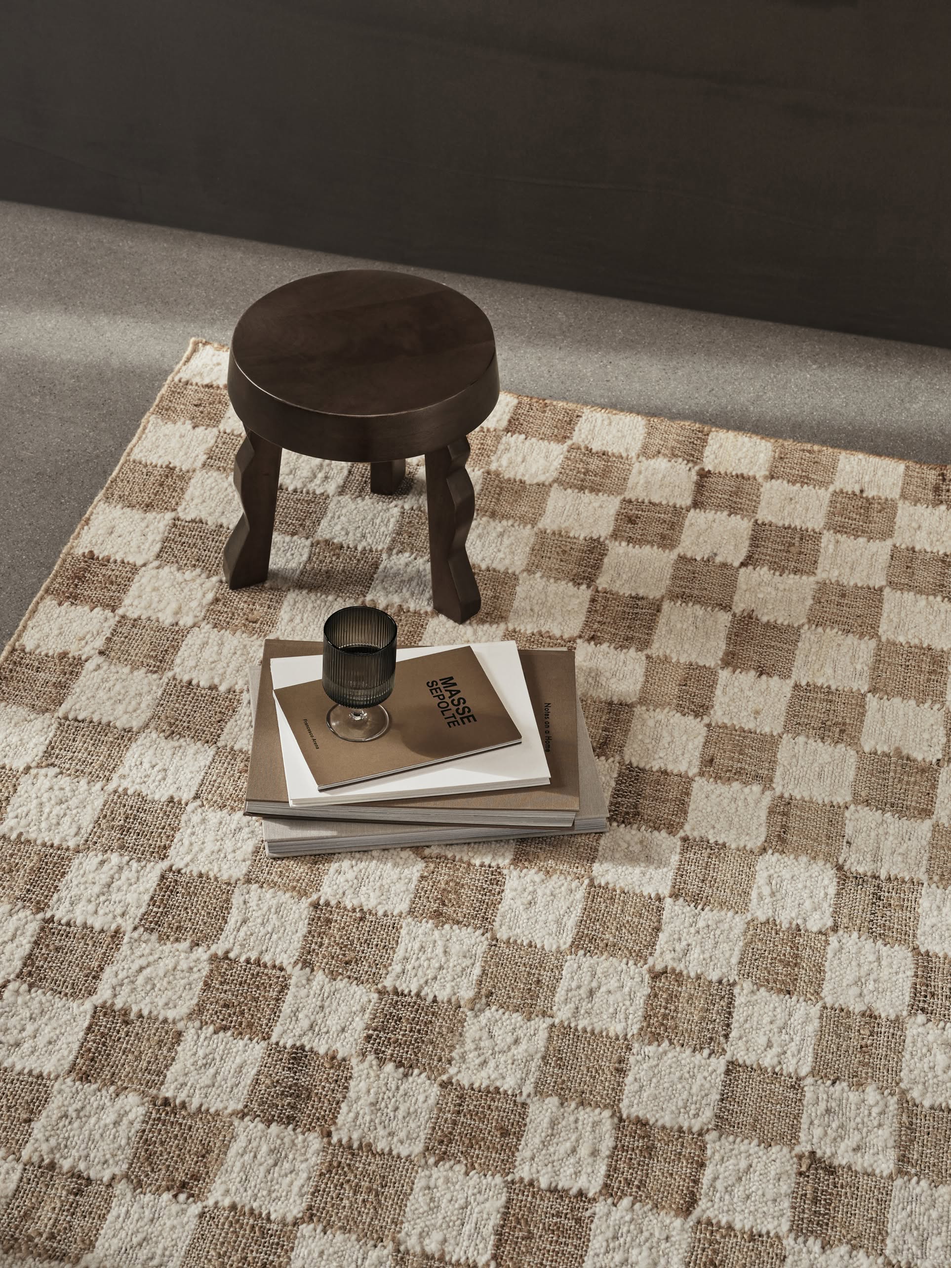 A small dark wooden stool sits on a gray floor beside the ferm LIVING Check Wool Jute Rug (200x300), its timeless beige-and-white check pattern enhancing the space. Stacked books and a glass cup add charm to the serene setting.