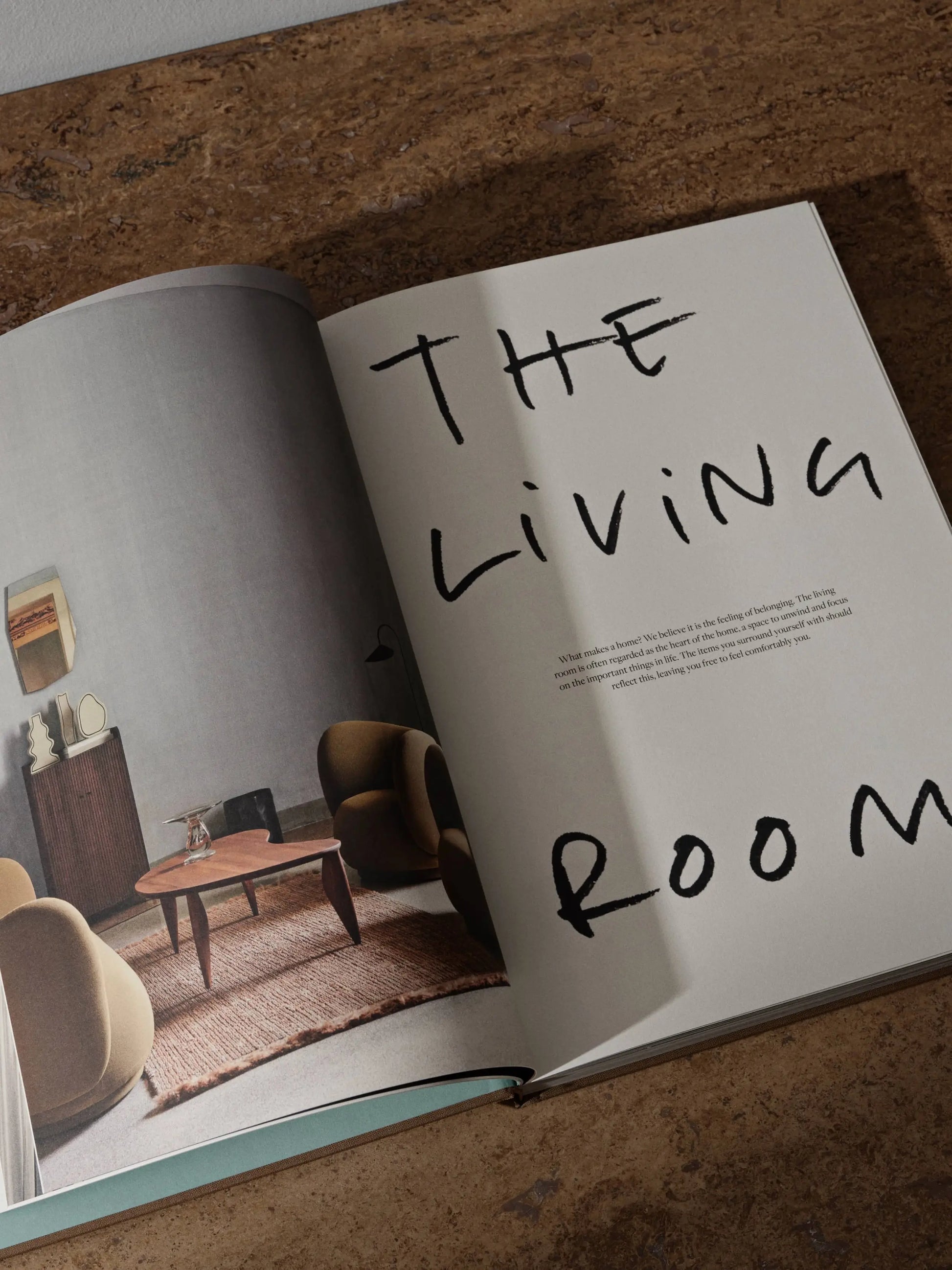 The ferm LIVING Coffee Table Book 2023 by ferm LIVING is open on a brown surface, featuring a modern living room photo on the left and large handwritten “THE LIVING ROOM” text over minimal text on the right.