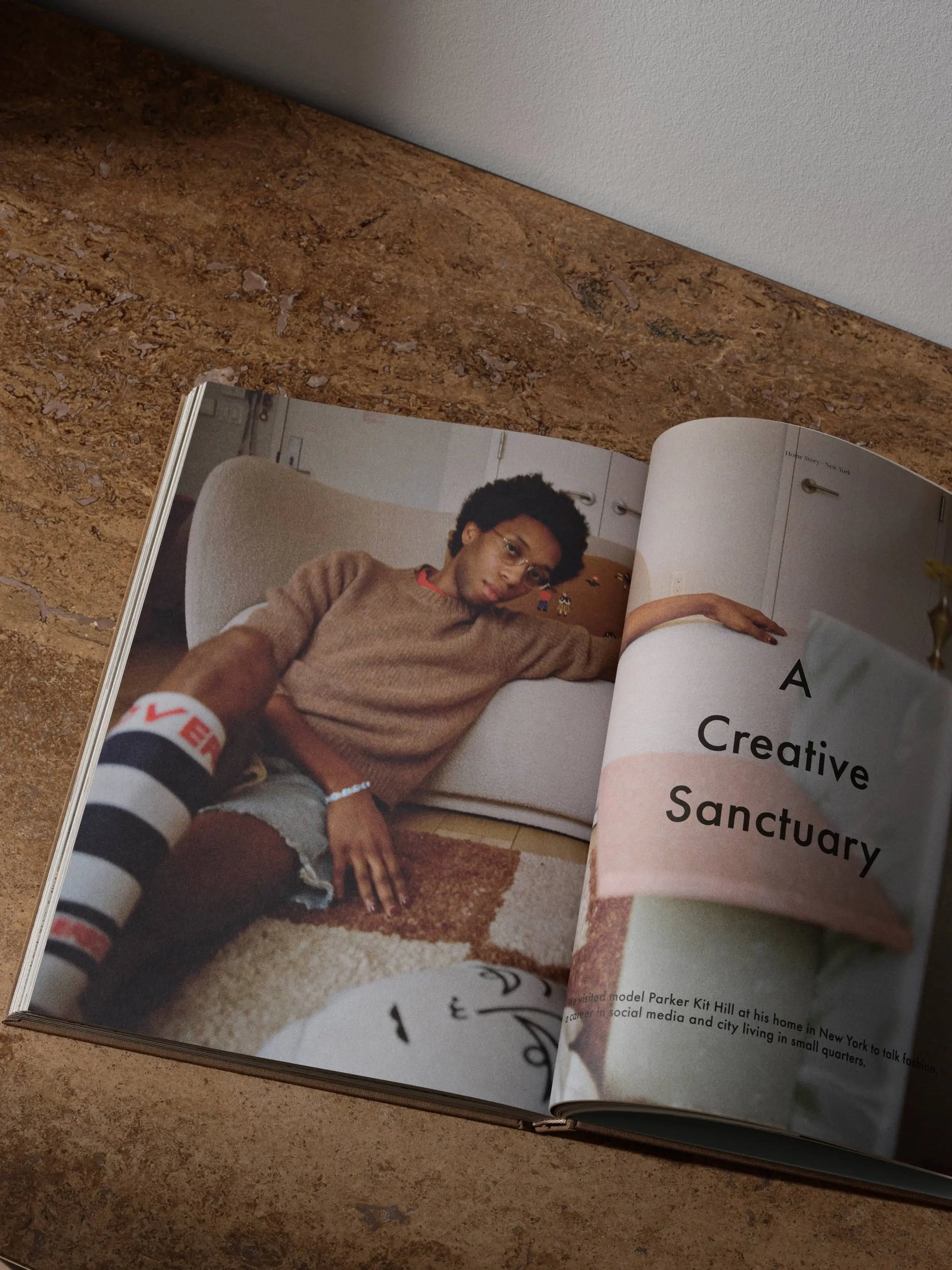 The ferm LIVING Coffee Table Book 2023 rests open on a brown surface, showing a young person on a sofa and the headline “A Creative Sanctuary,” reflecting ferm LIVING’s design ethos, with partial text and a pillow in the foreground.