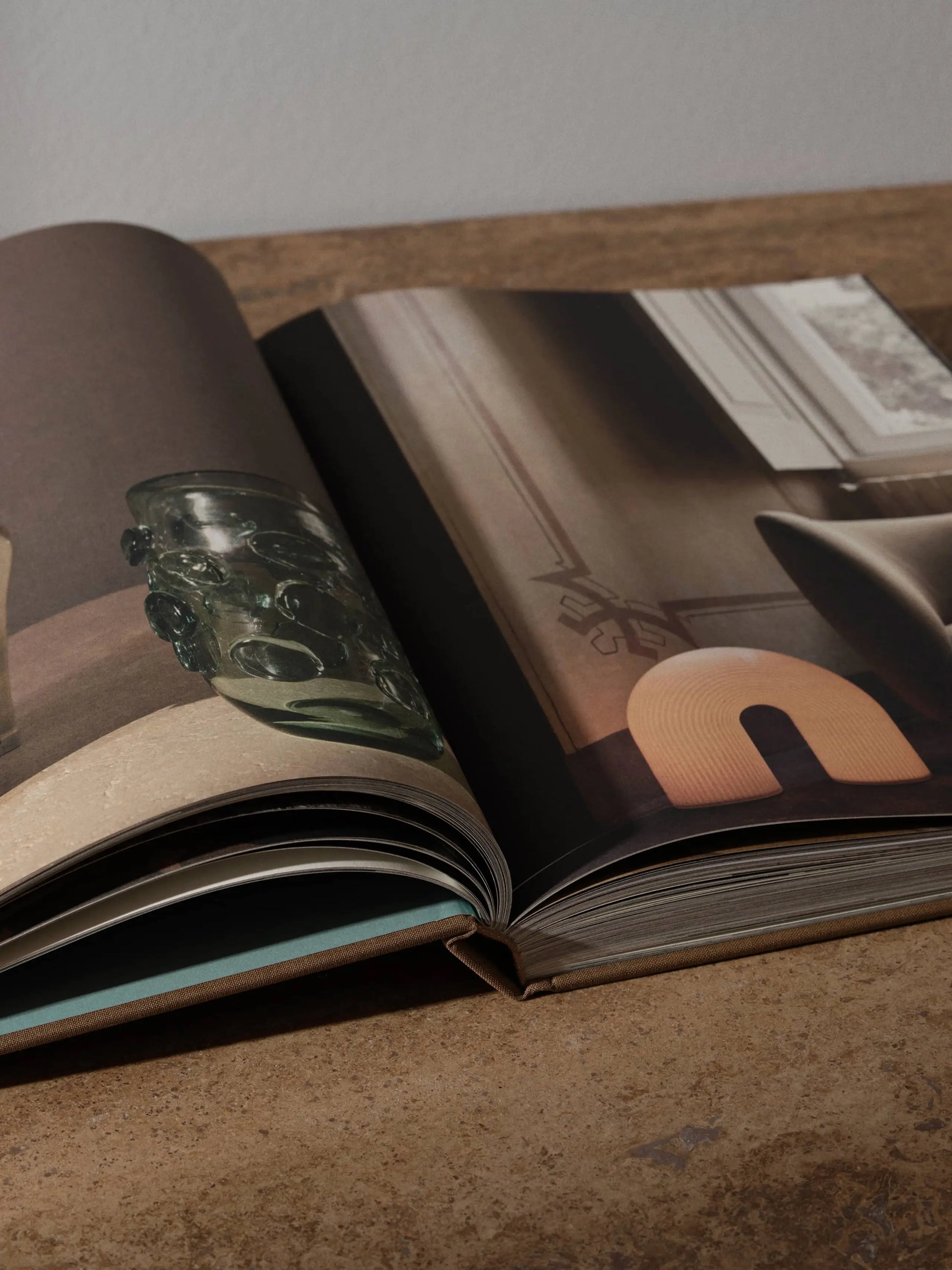 The ferm LIVING Coffee Table Book 2023 by ferm LIVING rests open on a wooden surface, showcasing photos of modern interiors—featuring a green glass vase and an arch-shaped lamp—perfect inspiration for any design project.