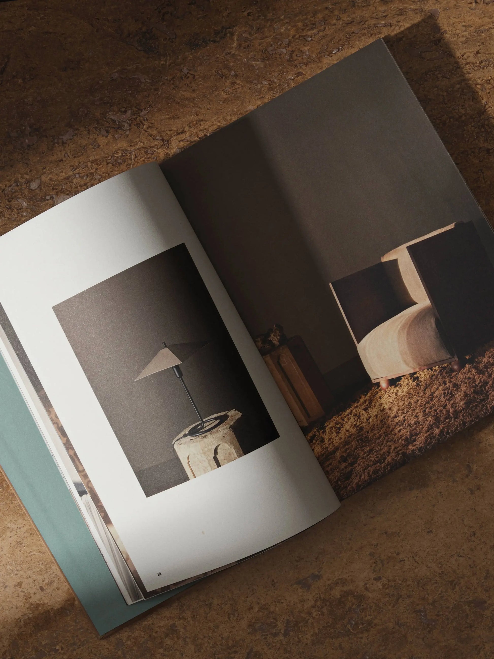 The ferm LIVING Coffee Table Book 2023 by ferm LIVING is open on a brown surface, featuring minimalist interior photos—a modern lamp on a stone table and a contemporary chair on a textured rug.