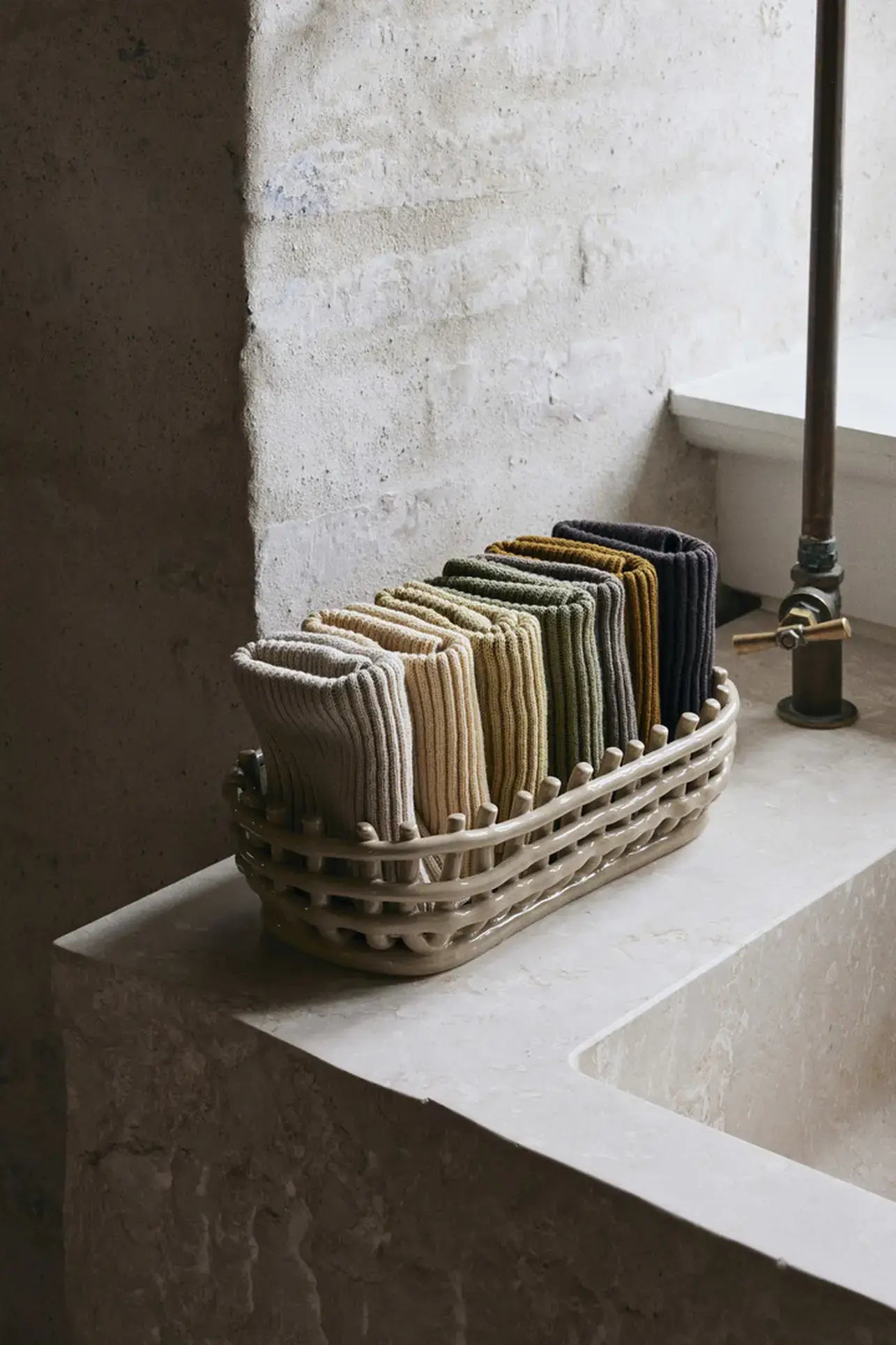 A woven basket on a stone sink holds neatly folded ferm LIVING Day Cloths, Set of 7—GOTS-certified organic cotton washcloths in beige, tan, green, and dark gray—beside a wall and metal faucet.