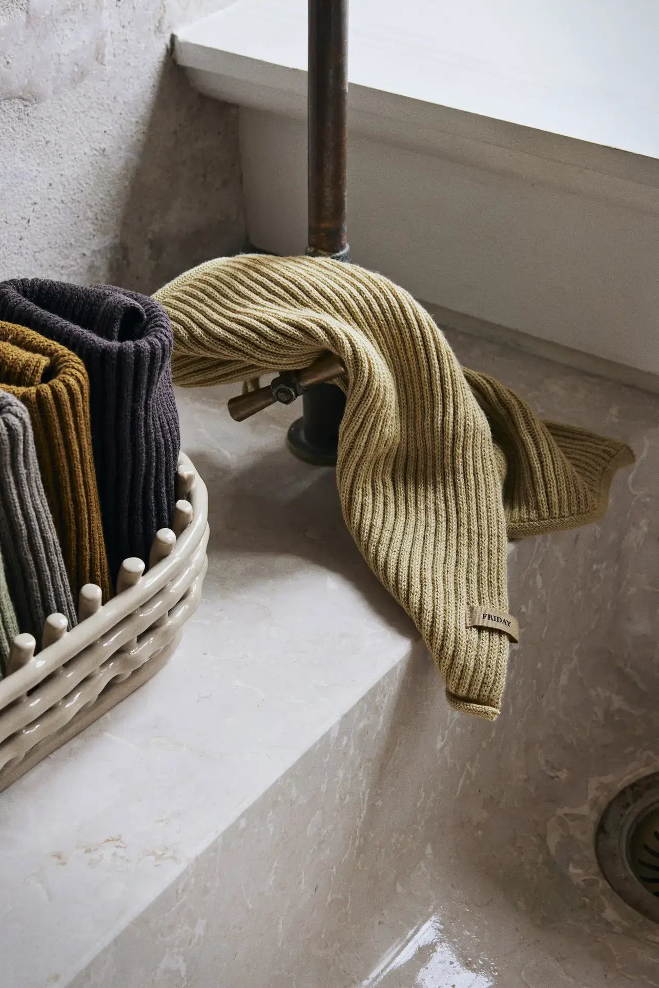 A set of ferm LIVING Day Cloths, including ribbed, mustard and dark neutral GOTS-certified towels, is draped over a marble sink next to a basket of folded cloths. An exposed pipe faucet rises above the scene.