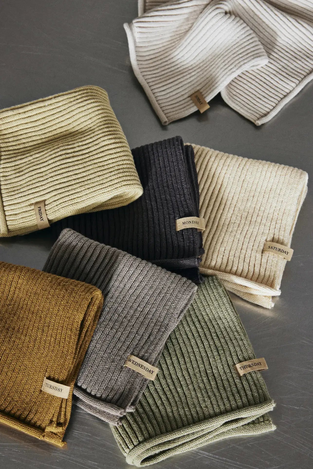 The ferm LIVING Day Cloths, Set of 7, are GOTS-certified organic cotton ribbed knit cloths, each labeled with a day of the week and neatly arranged on a gray surface.