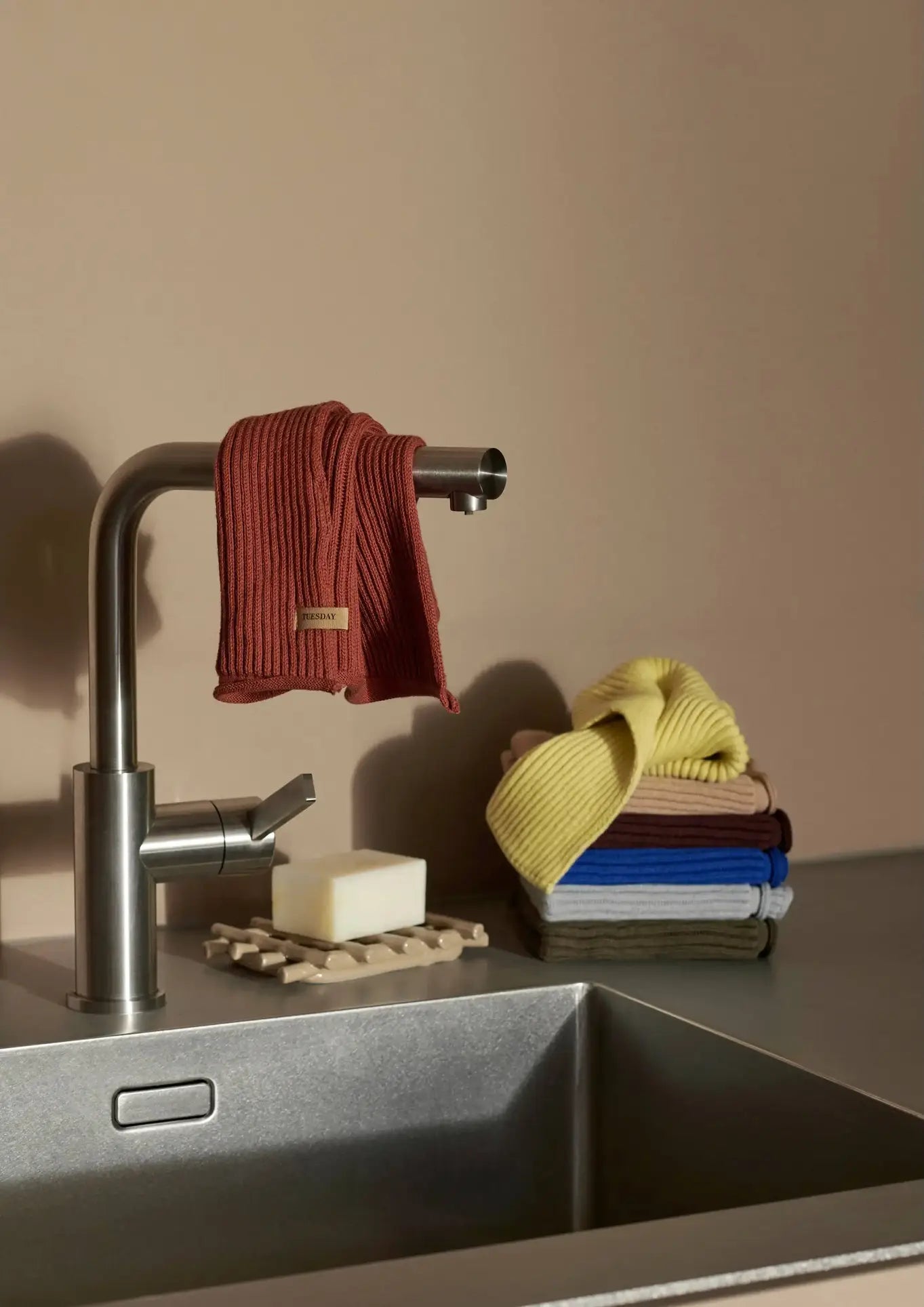 A modern kitchen sink with a metallic faucet, a red ribbed organic cotton dishcloth draped over the spout, a bar of soap on a tray, and a neat stack of ferm LIVING Day Cloths (Set of 7) by ferm LIVING on the counter.