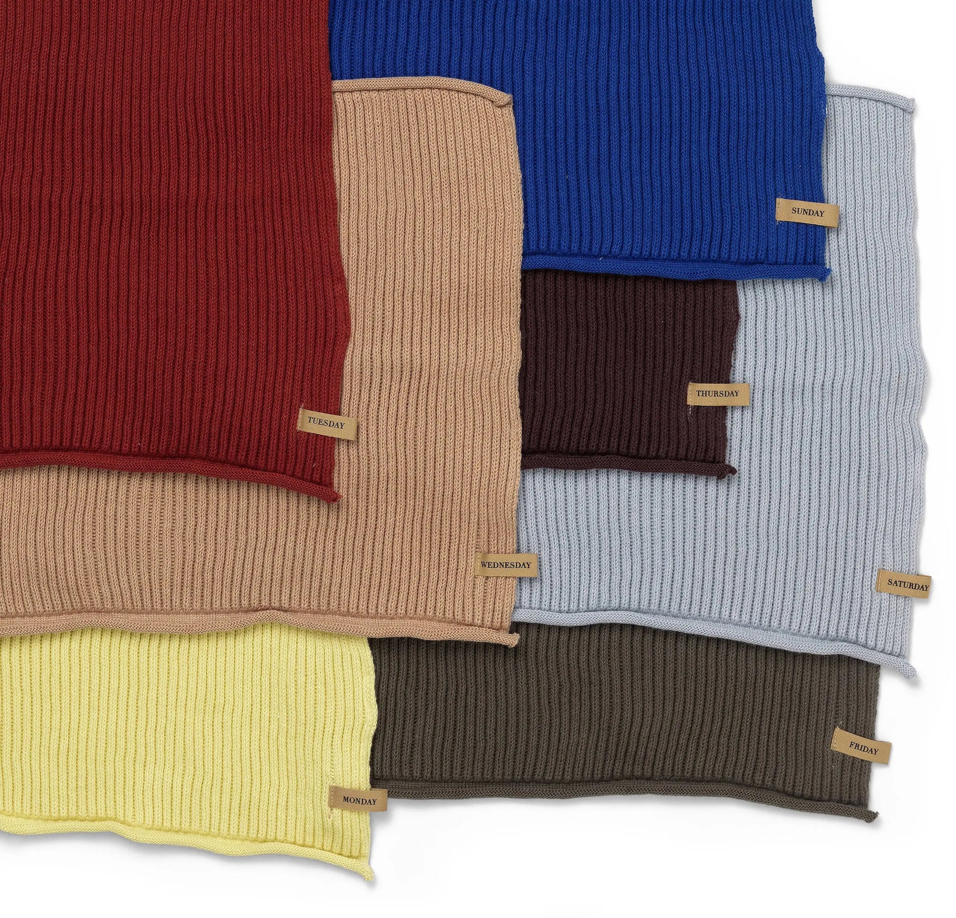 Seven GOTS-certified organic cotton ribbed knit cloths from the ferm LIVING Day Cloths Set, each labeled with a day of the week—including Monday, Thursday, Sunday, Friday, and Saturday—are shown overlapping in assorted colors.