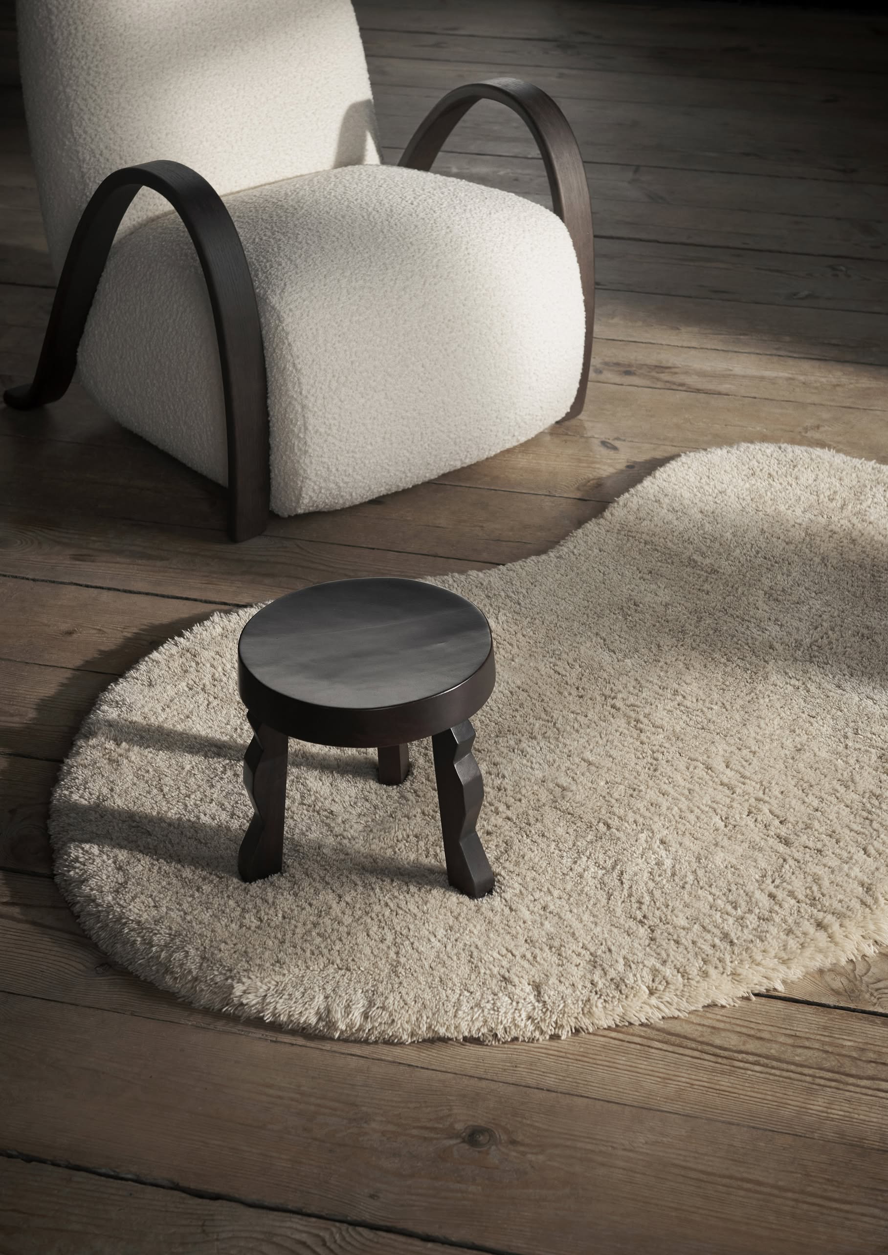 The ferm LIVING Faye Stool, a small round black accent, sits on a cream, irregular-shaped rug beside a modern white armchair with dark wooden arms atop wooden floorboards in a softly lit room.