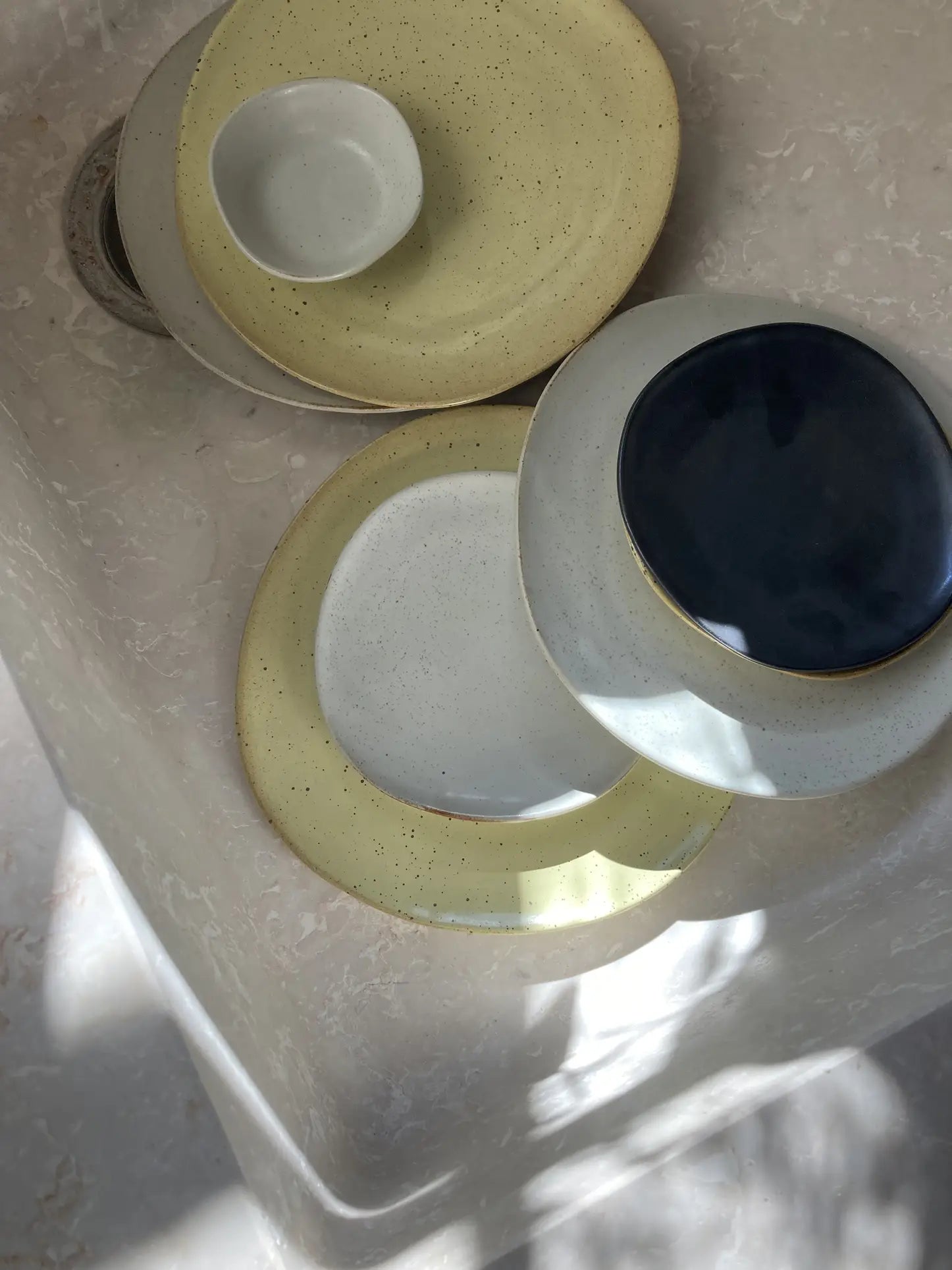 The ferm LIVING Flow Plate, Medium by ferm LIVING is stacked with matching ceramic tableware on marble, sunlight casting soft shadows over its modern semi-matte finish in muted tones of yellow, white, and blue.