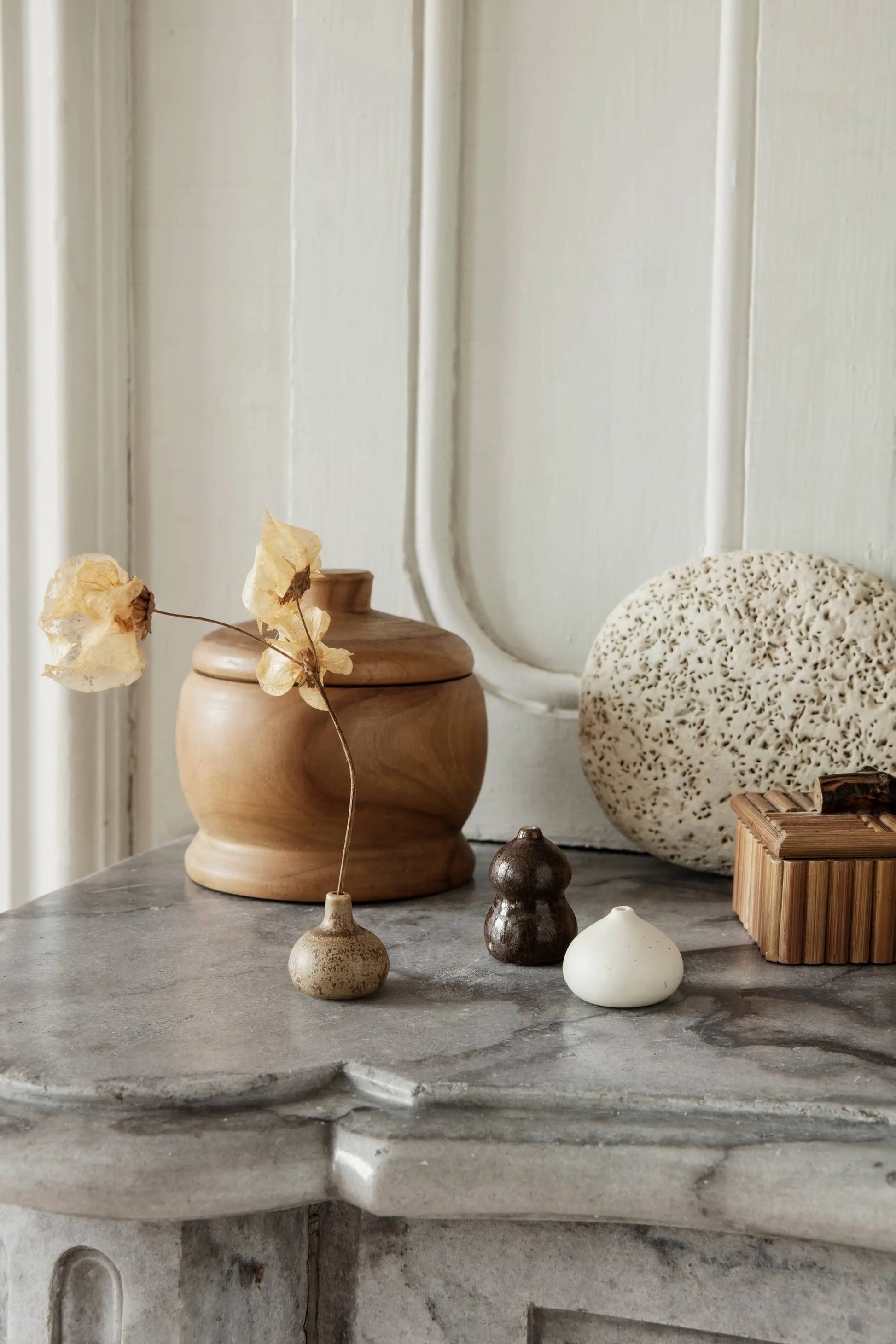 A minimalist still life on a gray surface features dried flowers in the ferm LIVING Komo Mini Vases, set of 3, with wooden containers, a round stone, and ceramic ornaments with reactive glazes against a white paneled wall.