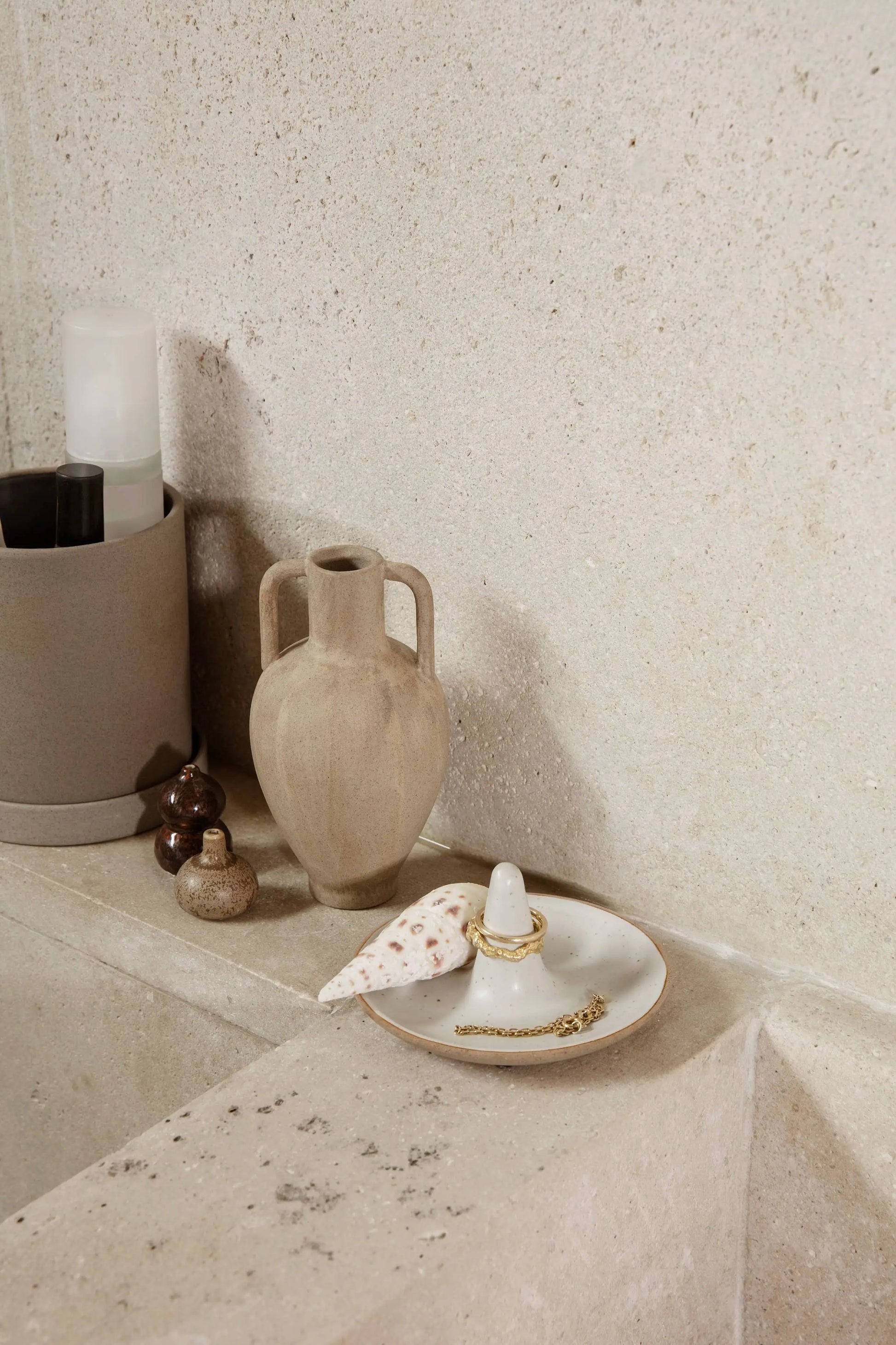 A beige stone bathroom countertop features the ferm LIVING Komo Mini Vases, Set of 3, a tall ceramic vase, a small brown bottle, a shell, gold jewelry on a ring holder, and toiletries in a round container against a textured stone wall.