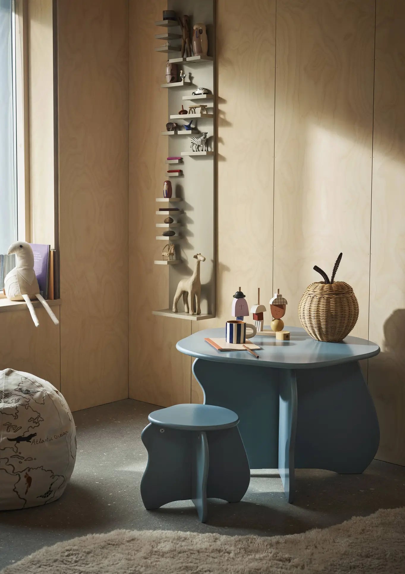 A cozy kids’ corner with a light blue table and stool, wooden animal figurines on wall shelves, a woven basket, window-side books, a plush pouf, and the ferm LIVING Milu Snack Set with hand-painted stripes on the rug.