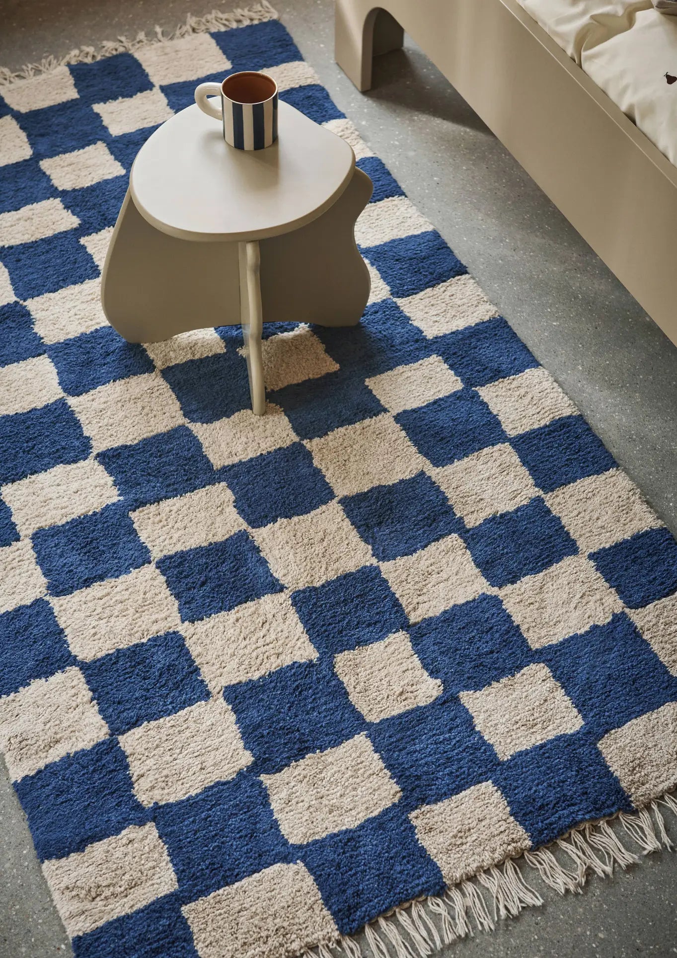A small round table with a ferm LIVING Milu Snack Set on top sits on a checkered blue and cream fringed rug next to a beige bed on a gray floor.