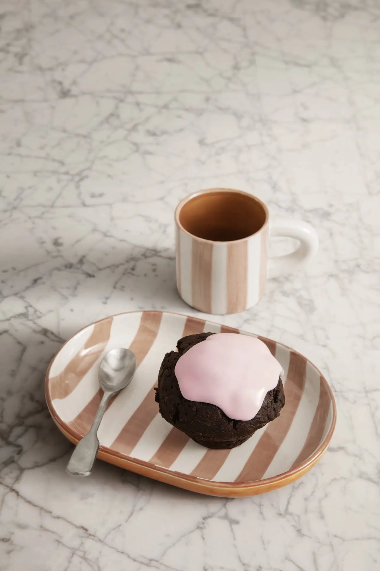 A glazed stoneware plate from the ferm LIVING Milu Snack Set holds a chocolate muffin with pink icing and a spoon on marble, while behind it sits the matching hand-painted mug by ferm LIVING filled with a dark beverage.