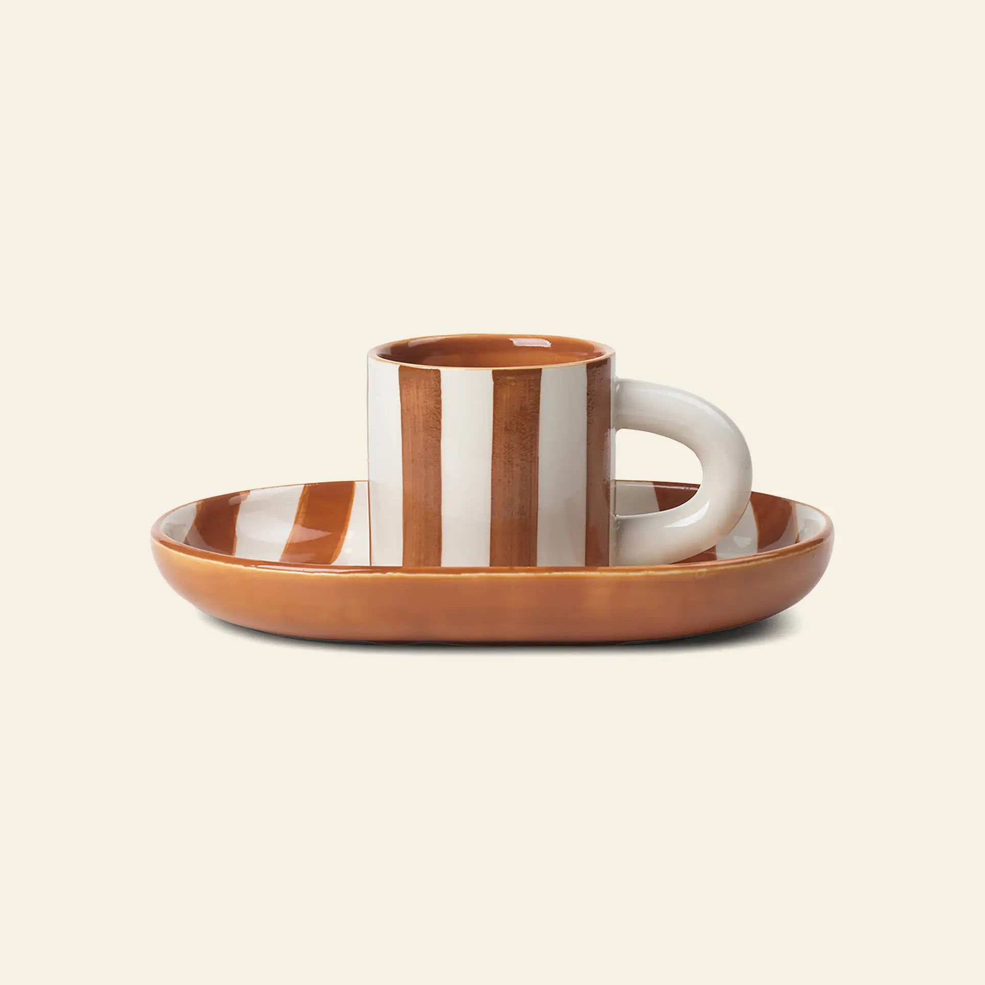 The ferm LIVING Milu Snack Set by ferm LIVING includes a glazed stoneware cup with vertical hand-painted brown and white stripes on a matching oval saucer with a brown rim, displayed on a plain beige background.