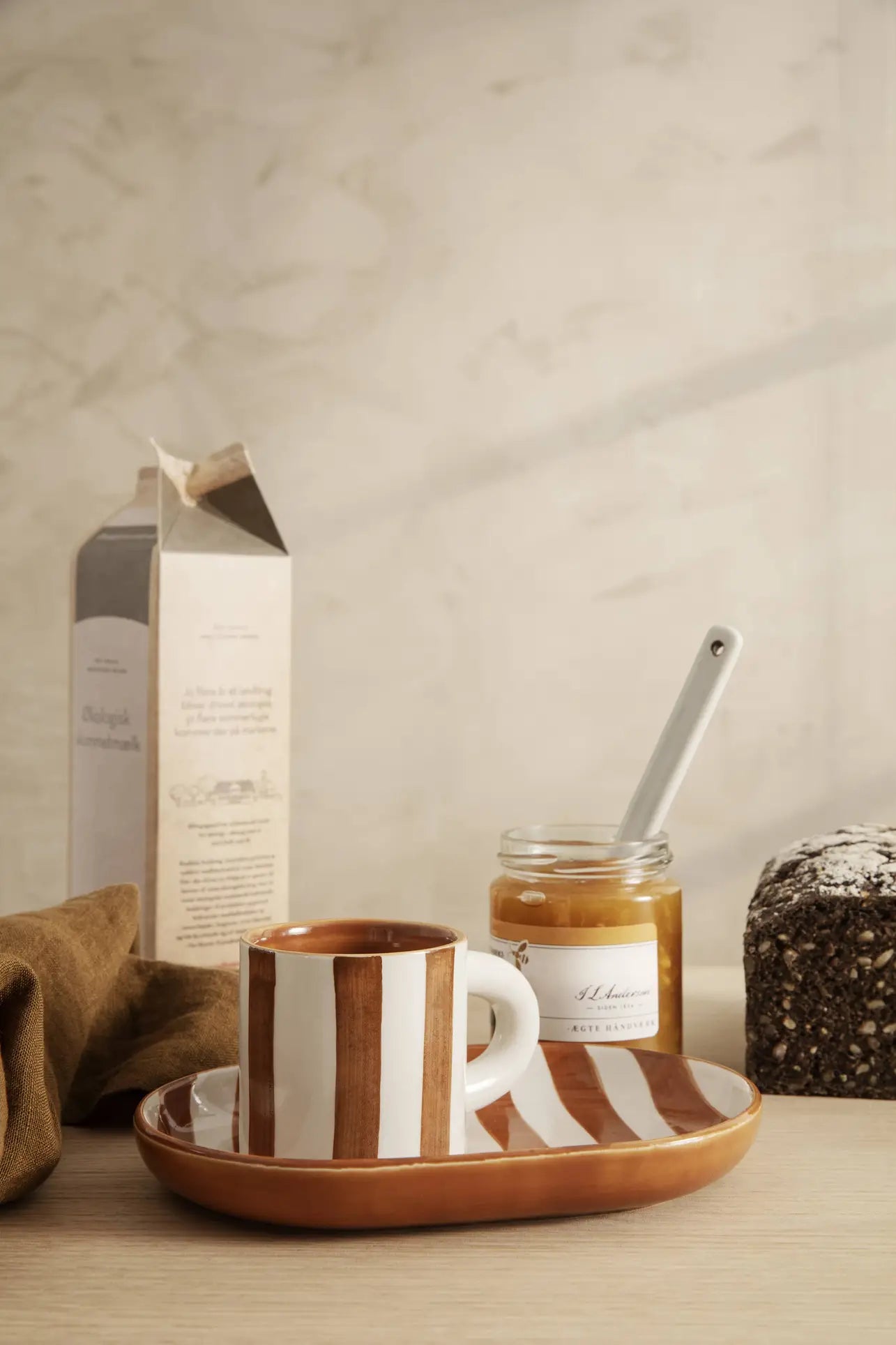 The ferm LIVING Milu Snack Set showcases a striped ceramic cup and saucer arranged on a wooden table with a honey jar, spoon, dark bread, brown napkin, and milk carton against a light background.