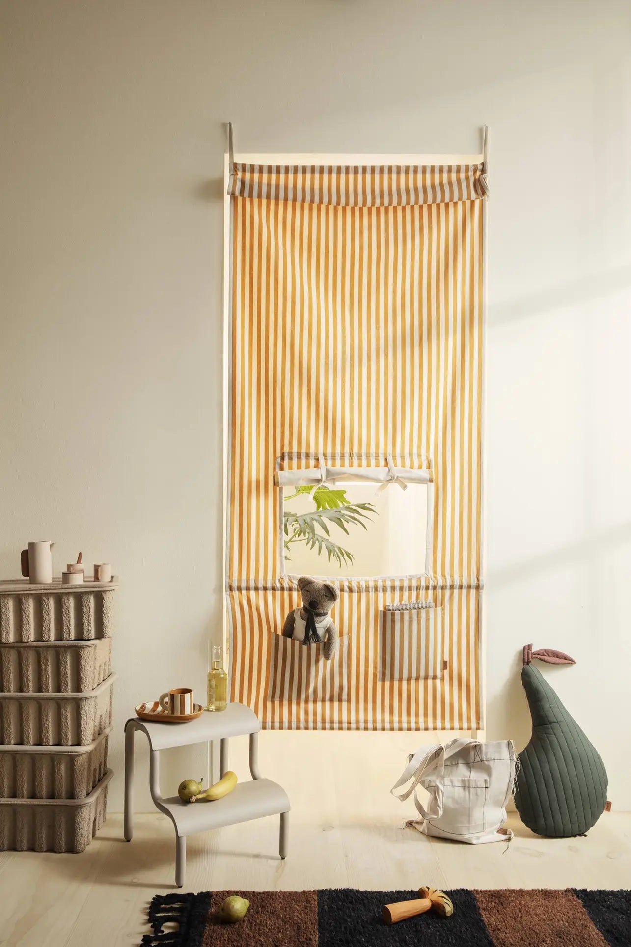 A sunlit room features a yellow and white striped hanging play tent, a stuffed elephant toy, a shelf with cups and books, a small table displaying the ferm LIVING Milu Snack Set, a green cushion, and a white bag on the floor.