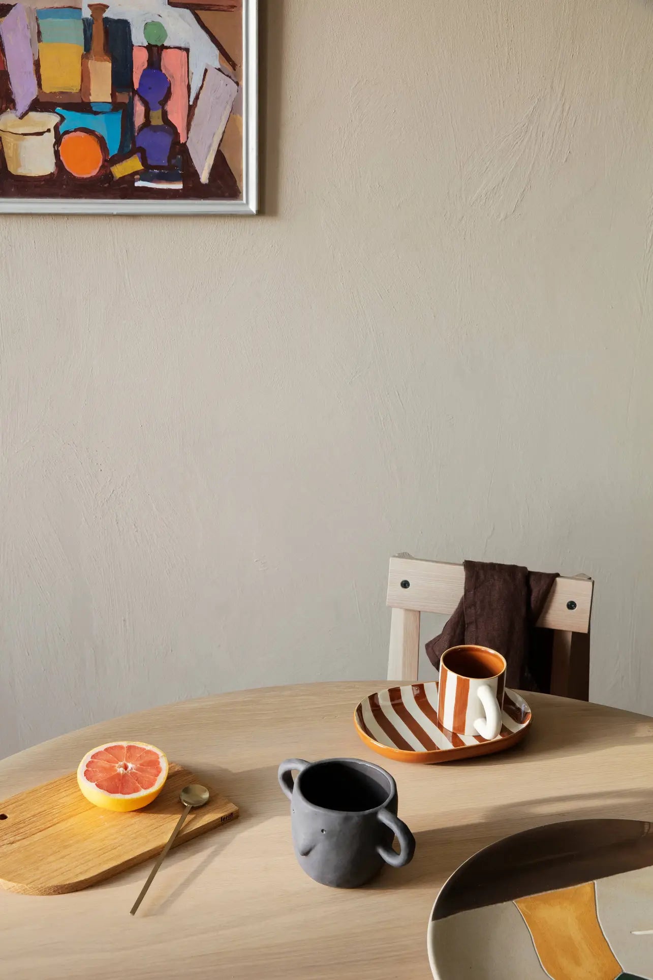 On a round wooden table sits the ferm LIVING Milu Snack Set—a cutting board with half a grapefruit and spoon, plus a dark gray mug. Nearby, a striped tray with cup and brown cloth on a chair. Abstract art decorates the beige wall behind.