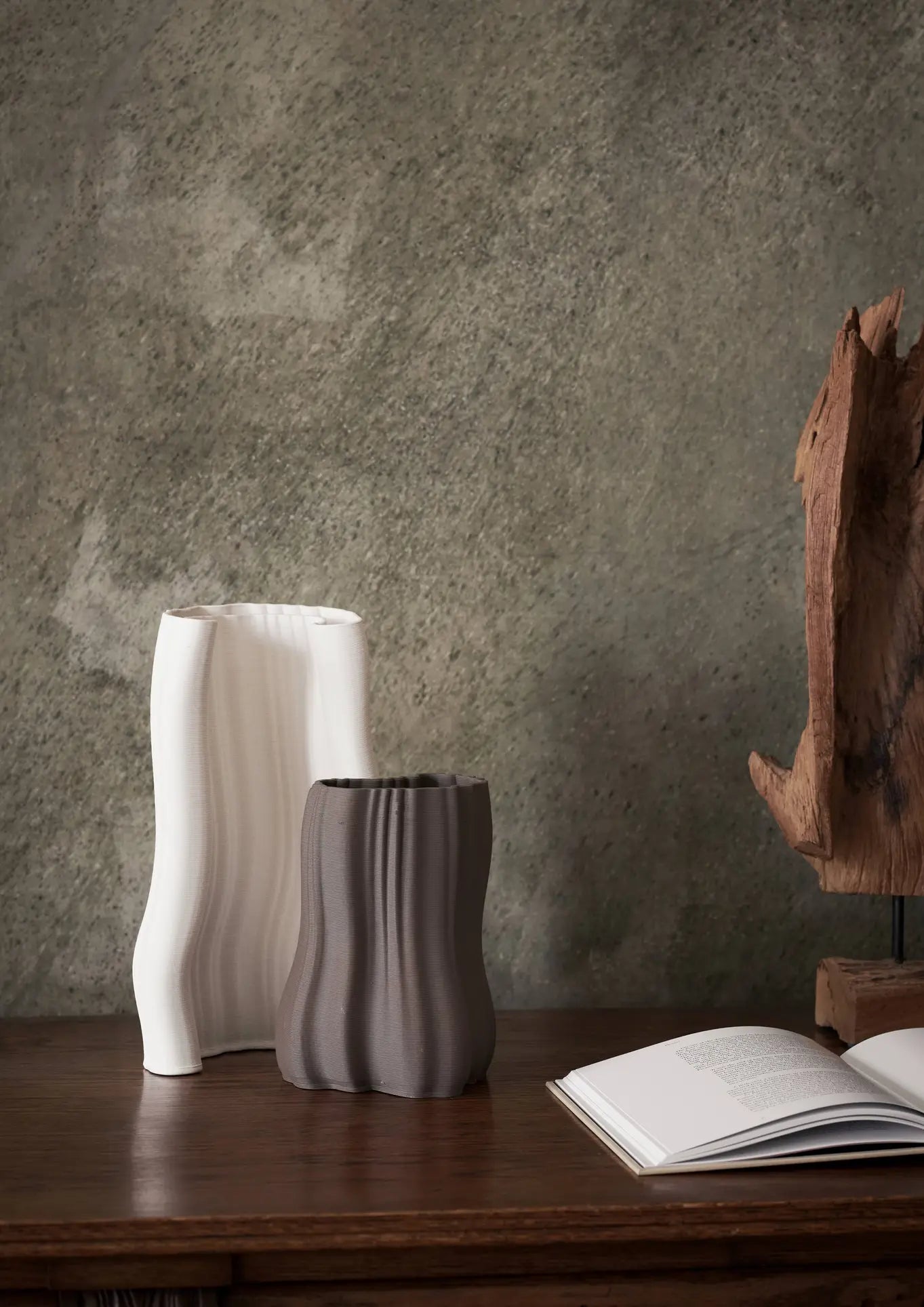 The ferm LIVING Moire Vase, Small sits with another ribbed ceramic vase on a wooden surface beside an open book and abstract wood sculpture, highlighting the artistry of 3D-printed clay against a textured gray wall backdrop.