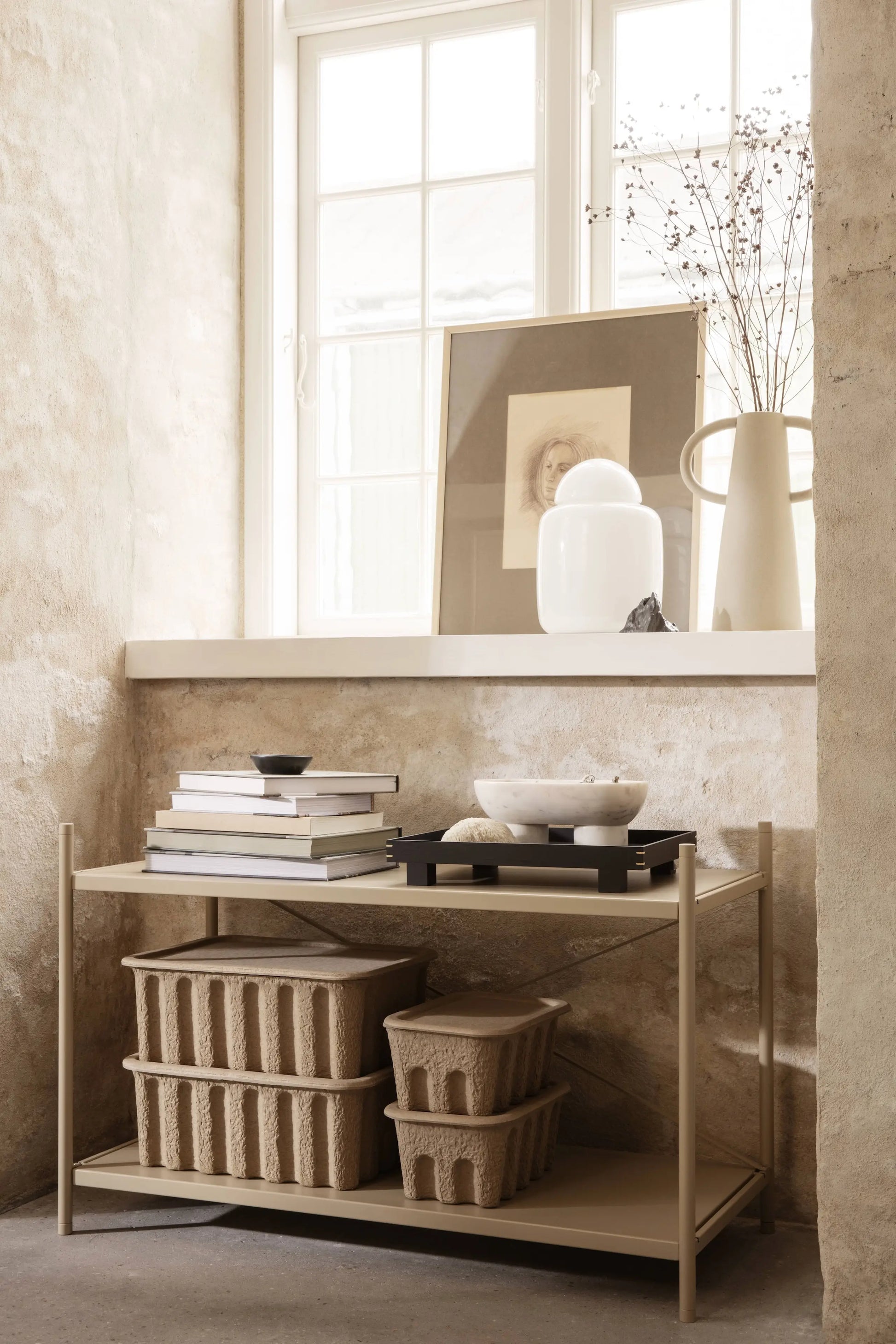 A minimalist beige shelf stands by a sunlit window, holding stacked books, decorative bowls, and a set of two ferm LIVING Paper Pulp Box Large. Above, a vase with dried branches and framed art rest on the windowsill against a textured wall.