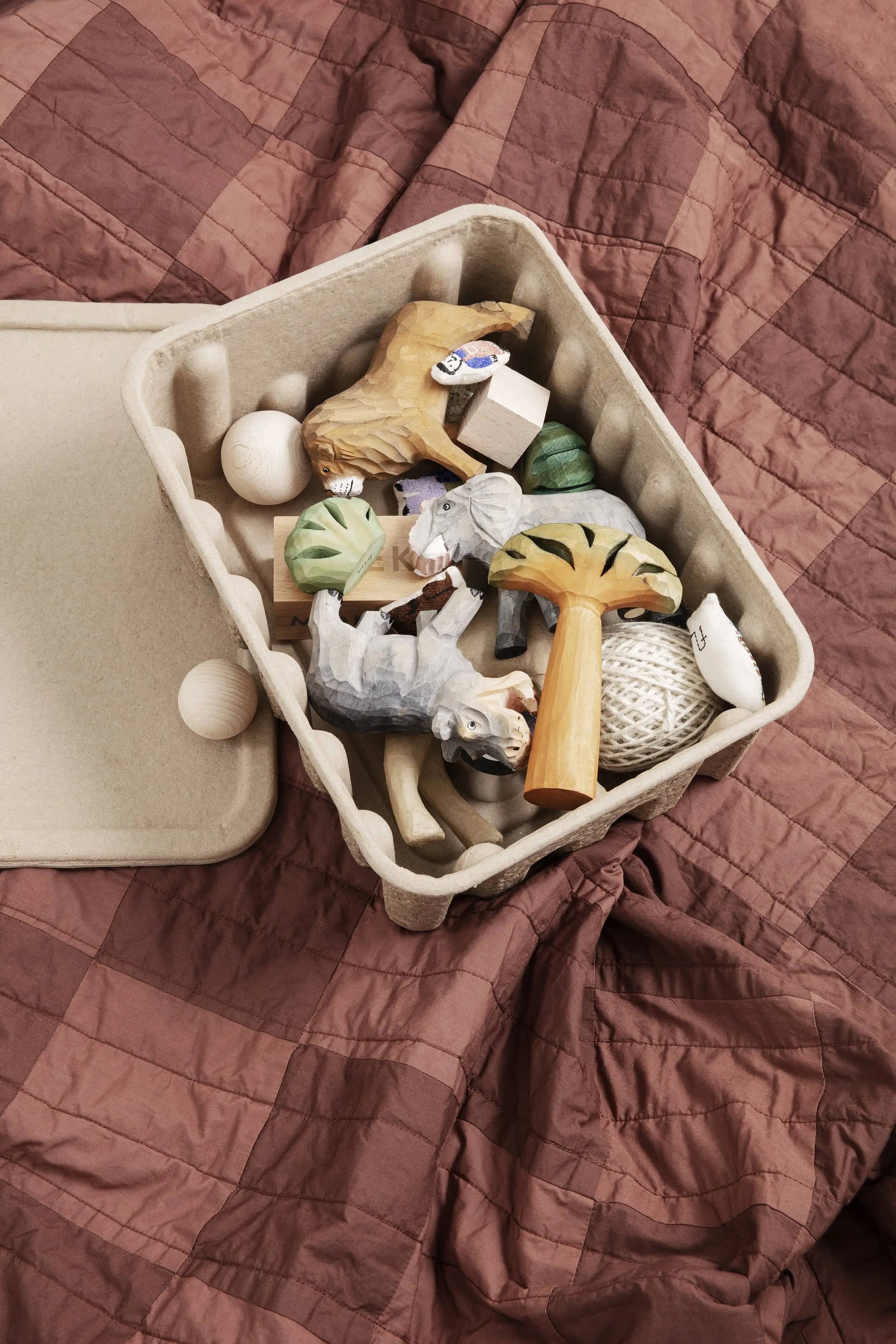 The ferm LIVING Paper Pulp Box Large, Set of 2, in beige is partially open and filled with wooden toys and textured balls, sitting atop a crumpled mauve quilted blanket—a stylish and eco-friendly storage solution from ferm LIVING.