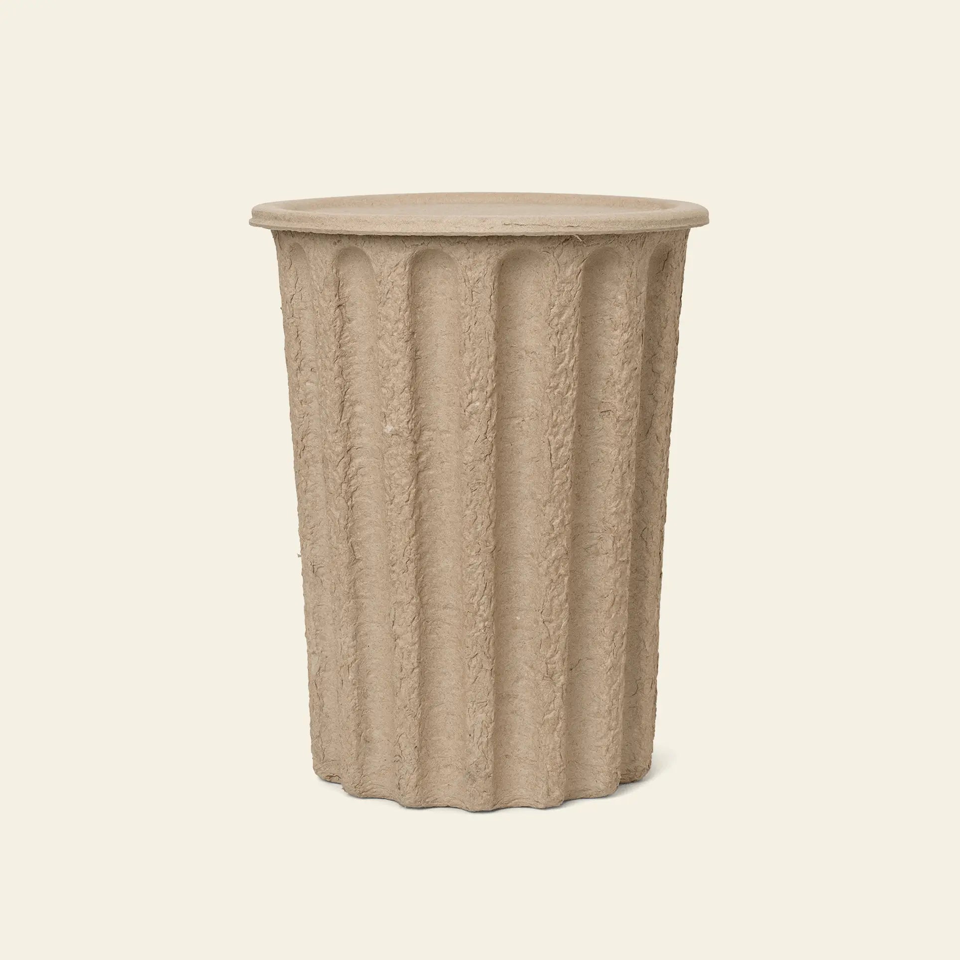 The ferm LIVING Paper Pulp Paper Bin features a cylindrical, beige, fluted design made from recycled paper pulp with a textured surface and flat top, echoing classical columns, set against a plain light background.
