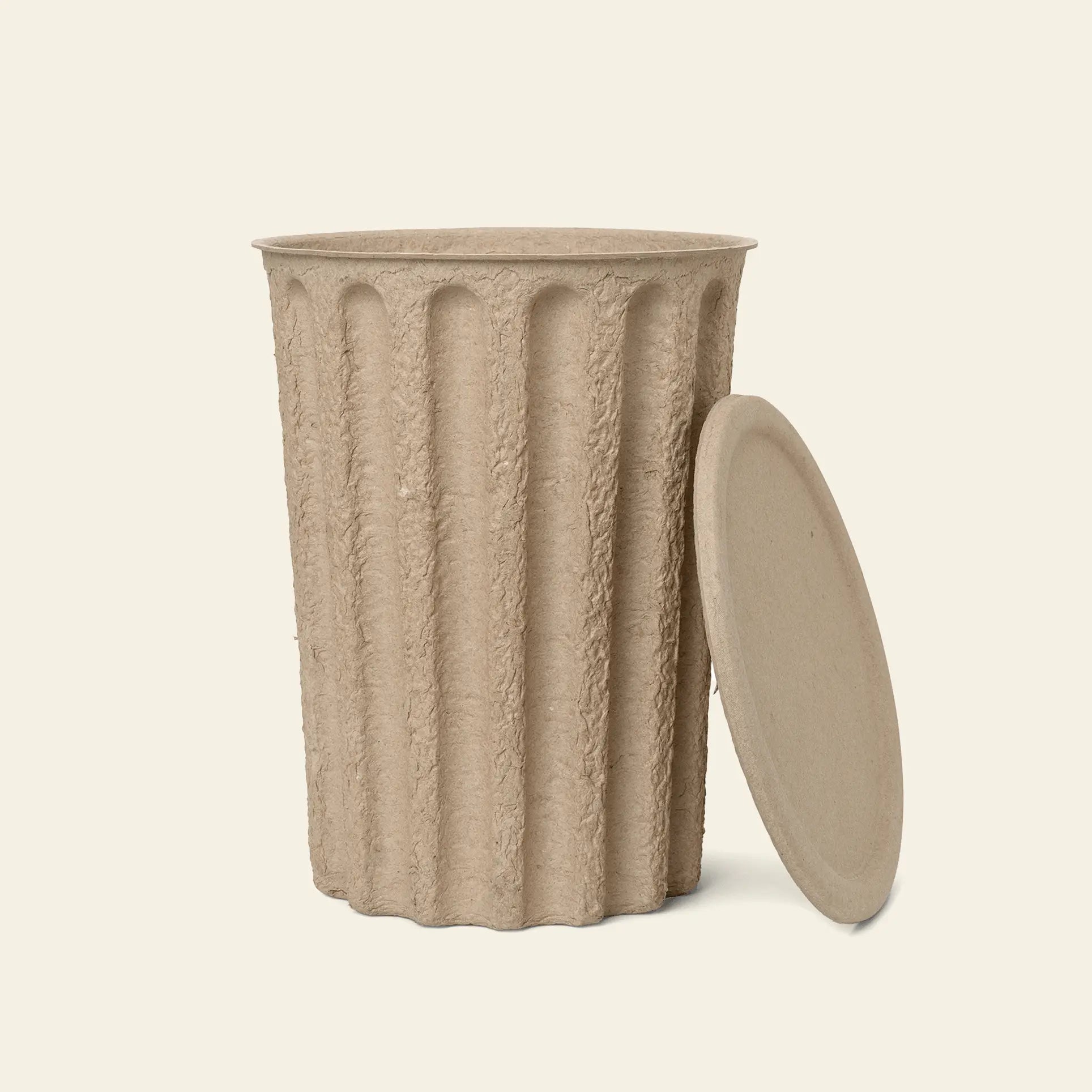 The ferm LIVING Paper Pulp Paper Bin is a textured beige cylinder with vertical ridges and a flat lid, both made from recycled paper pulp, shown against a plain light background.