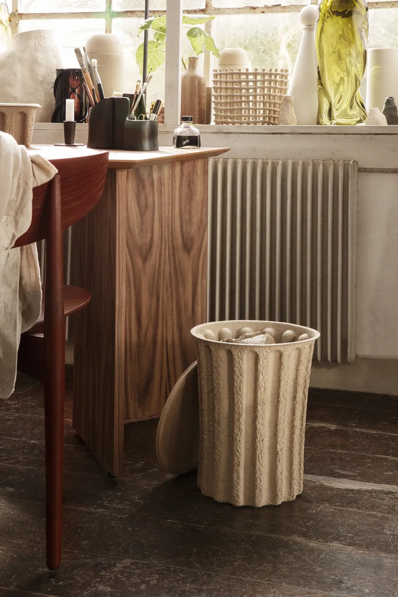 A neutral-toned workspace features a wooden desk, red chair, and the ferm LIVING Paper Pulp Paper Bin by ferm LIVING on the floor. Ceramics, plants, and glass objects line the windowsill, bathed in soft natural light.
