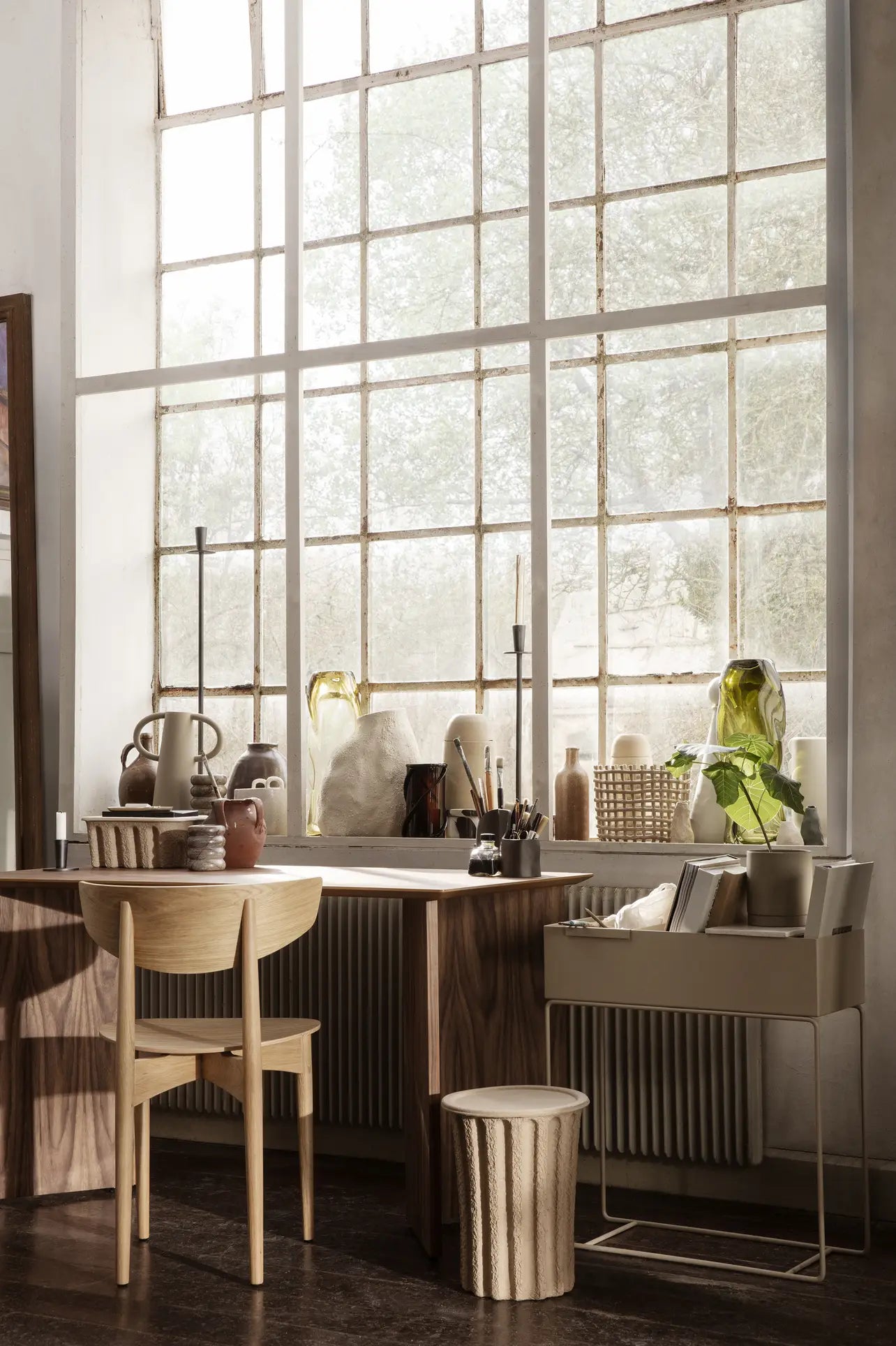 A sunlit workspace with a wooden desk and chair sits by large windows. Ceramics, vases, plants, and the ferm LIVING Paper Pulp Paper Bin by ferm LIVING decorate the area, creating a cozy, artistic vibe.