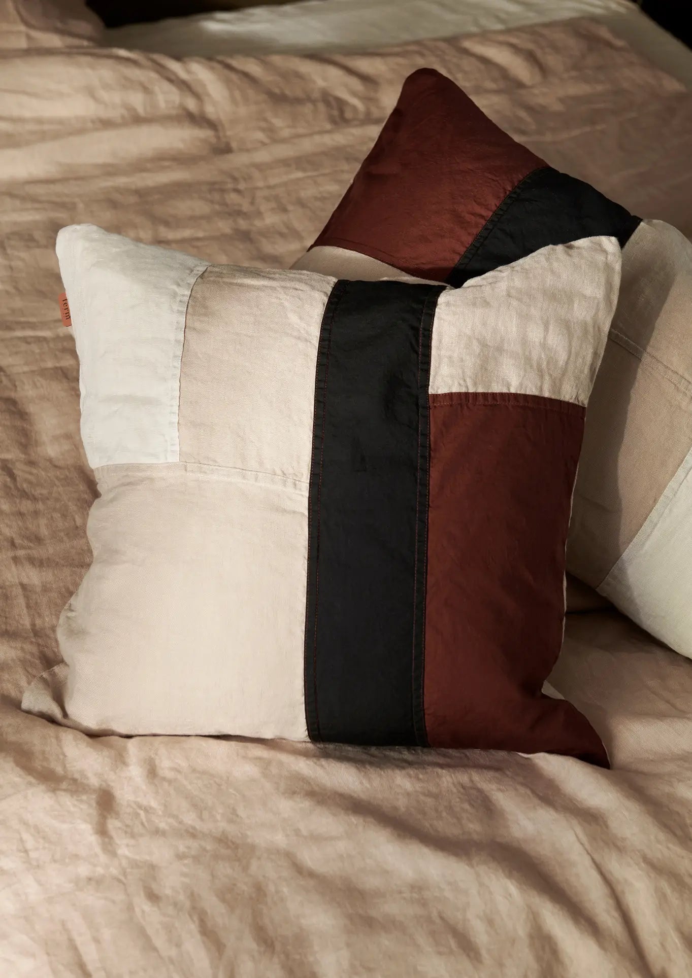 Two ferm LIVING Part Cushions with linen patchwork in beige, black, and brown sit on a neatly made beige bed, creating a cozy and minimalist look.