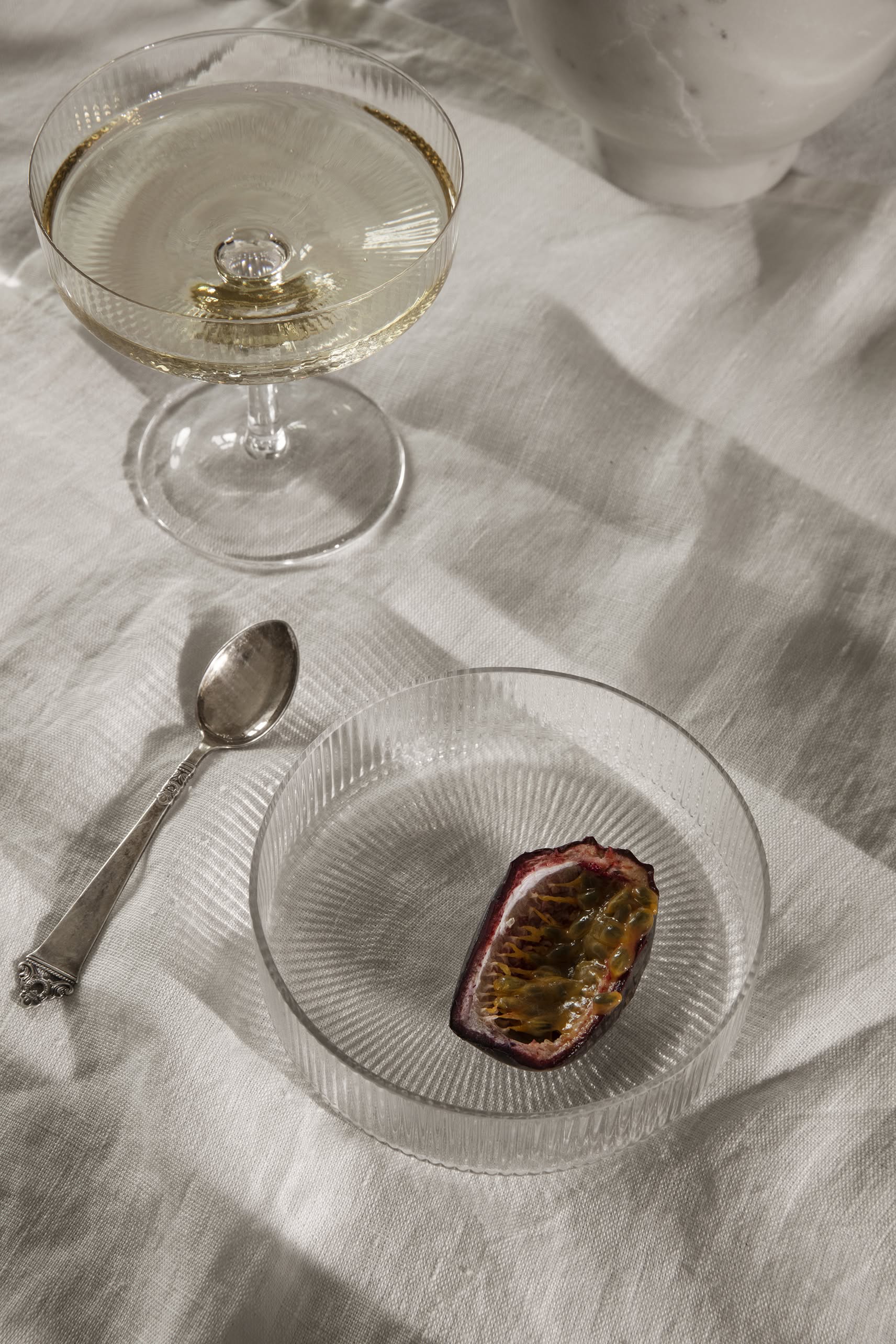 A mouth-blown coupe filled with a clear drink sits on a white textured cloth next to the ferm LIVING Ripple Serving Bowls, Set of 4, one holding half a passion fruit and a silver spoon.