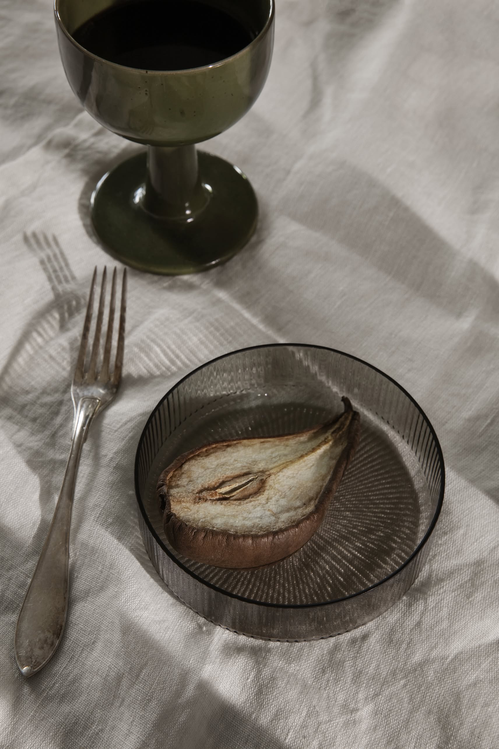 A sliced, dried pear sits on a ferm LIVING Ripple Serving Bowl beside a silver fork and a dark glass goblet, all elegantly arranged on a textured surface.