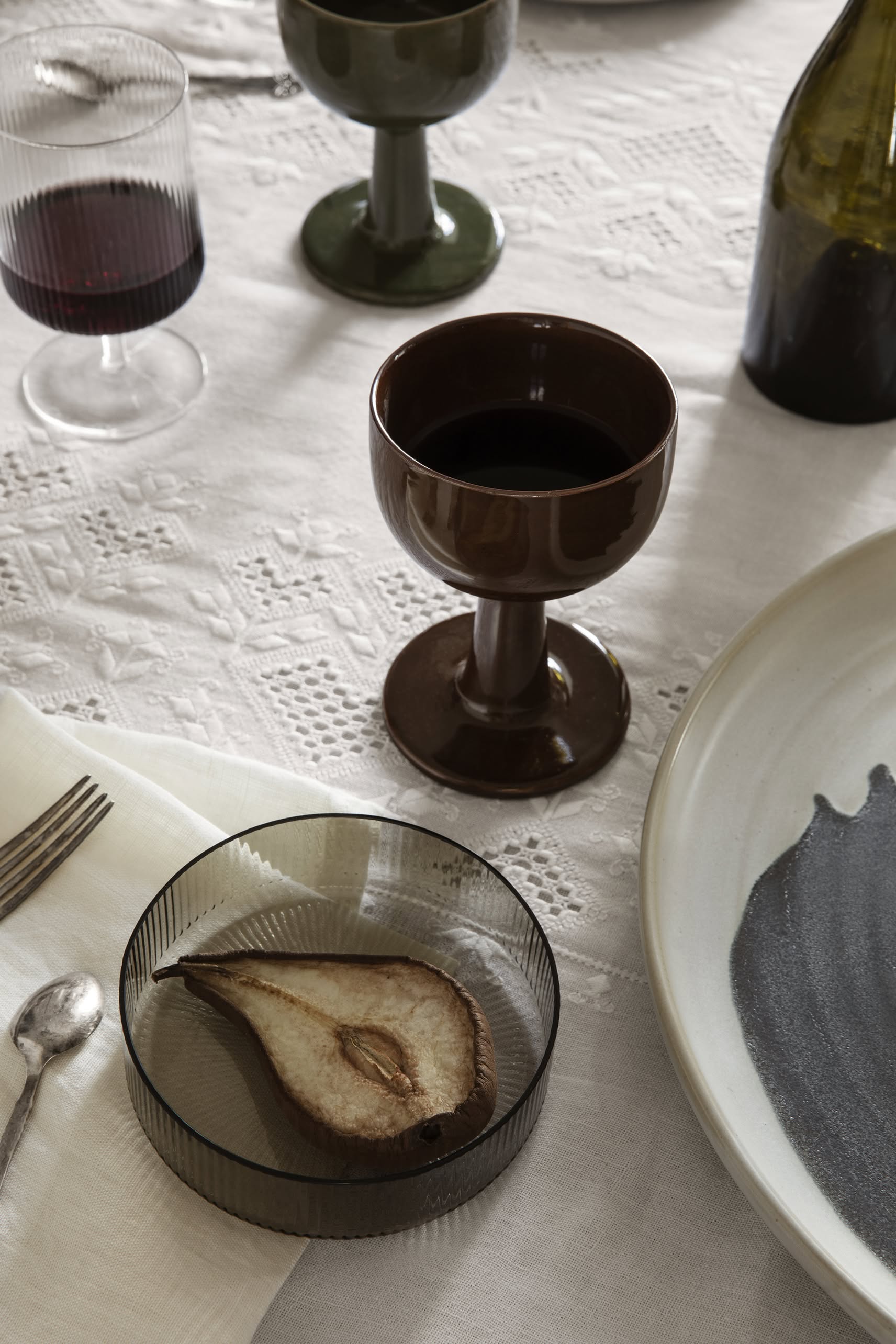 A table set with a mouth-blown glass of red wine, brown goblet, empty bottle, cutlery, napkin, and a dried pear slice in a ferm LIVING Ripple Serving Bowl from the Set of 4 on a white embroidered tablecloth.