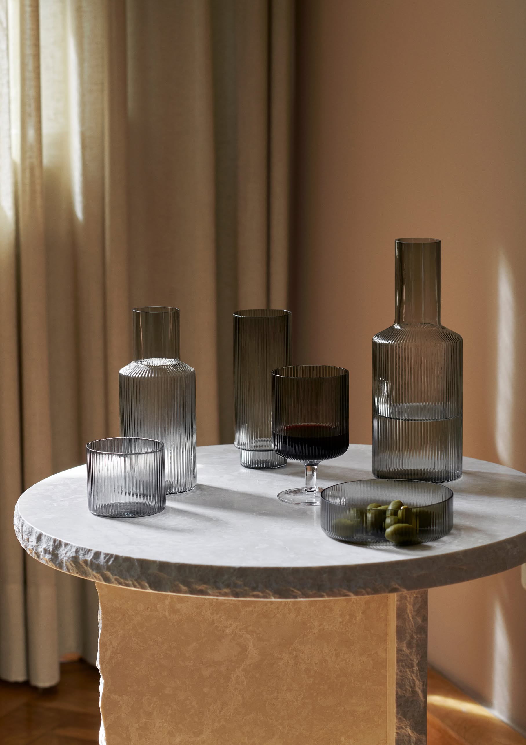 A round marble table displays ribbed, mouth-blown glassware and ferm LIVING Ripple Serving Bowls, Set of 4 by ferm LIVING, filled with green olives. Sunlight filters through beige curtains in the background.