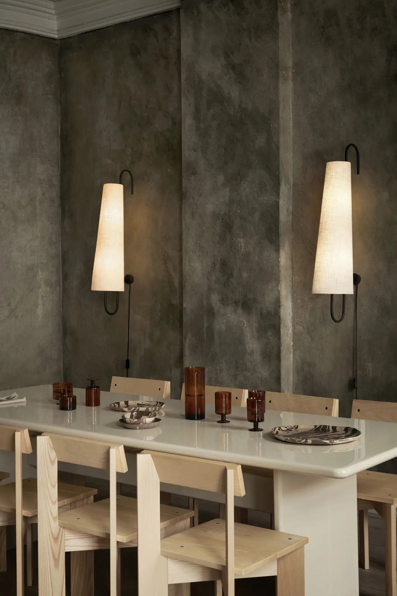 A modern dining area features a rectangular white table, light wooden chairs, brown glassware, and ferm LIVING Ryu Platters (18.5cm) beneath two large cone-shaped wall lamps against textured gray walls.