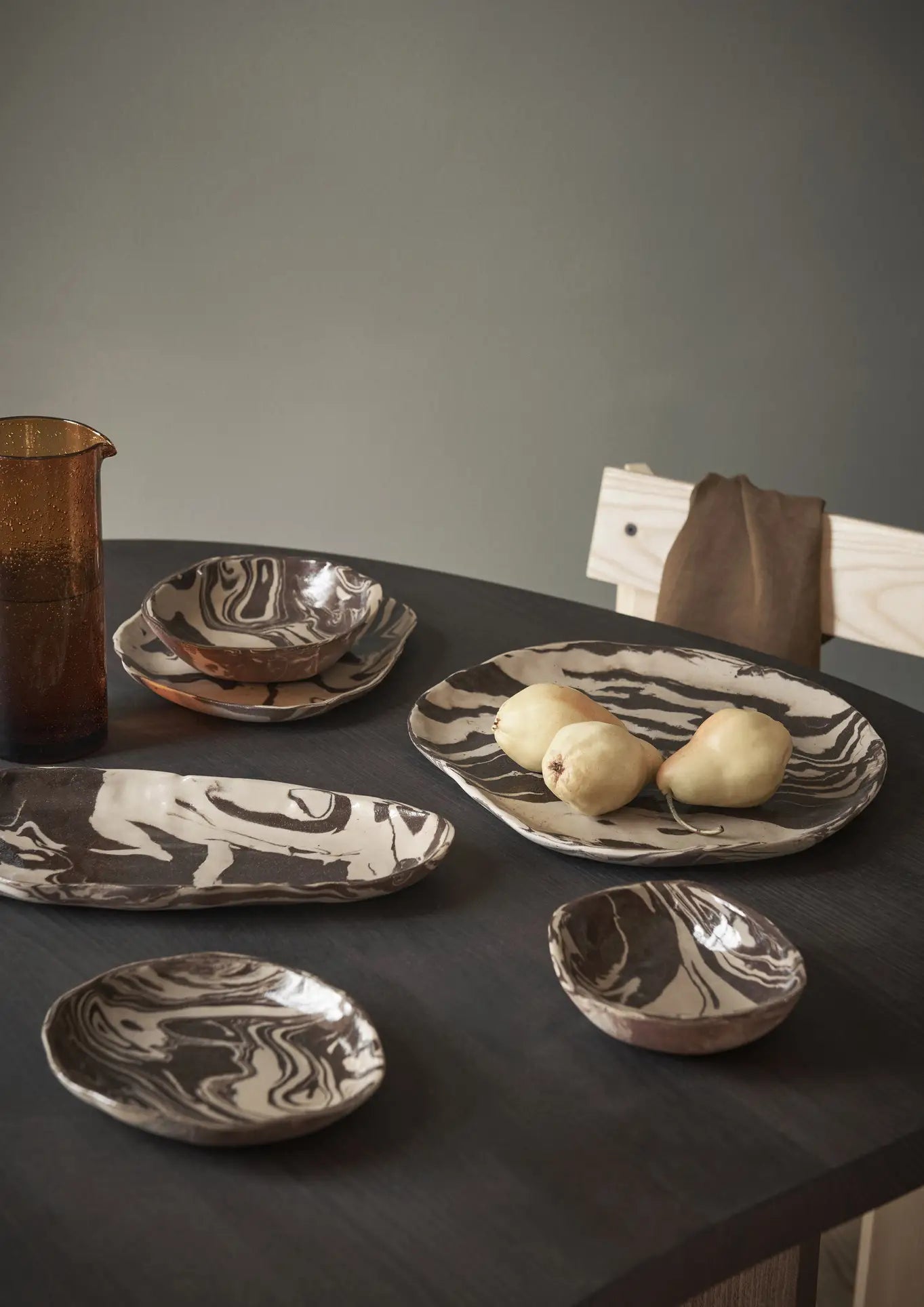 A dark table features the ferm LIVING Ryu Platter, 18.5cm, displaying marbled ceramics in neutral tones—some with yellow pears—plus a brown glass pitcher that brings warmth to the modern, minimalist space.