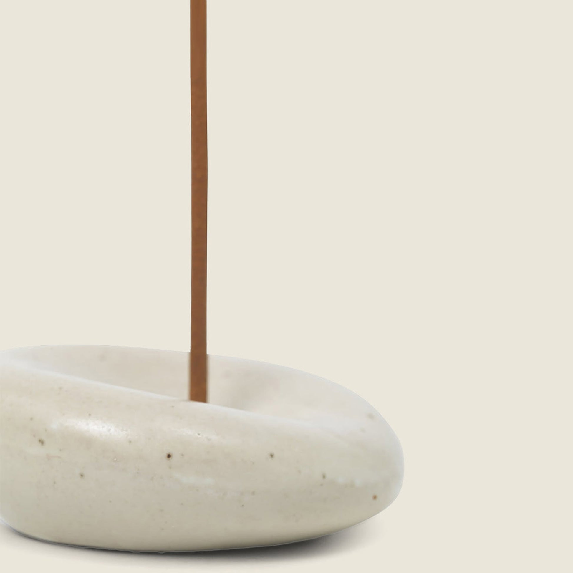 A brown sandalwood incense stick stands in the smooth, round ferm LIVING Sense Incense Holder Gift Set by ferm LIVING, its white ceramic holder gently dipped at the center and set against a plain beige background.